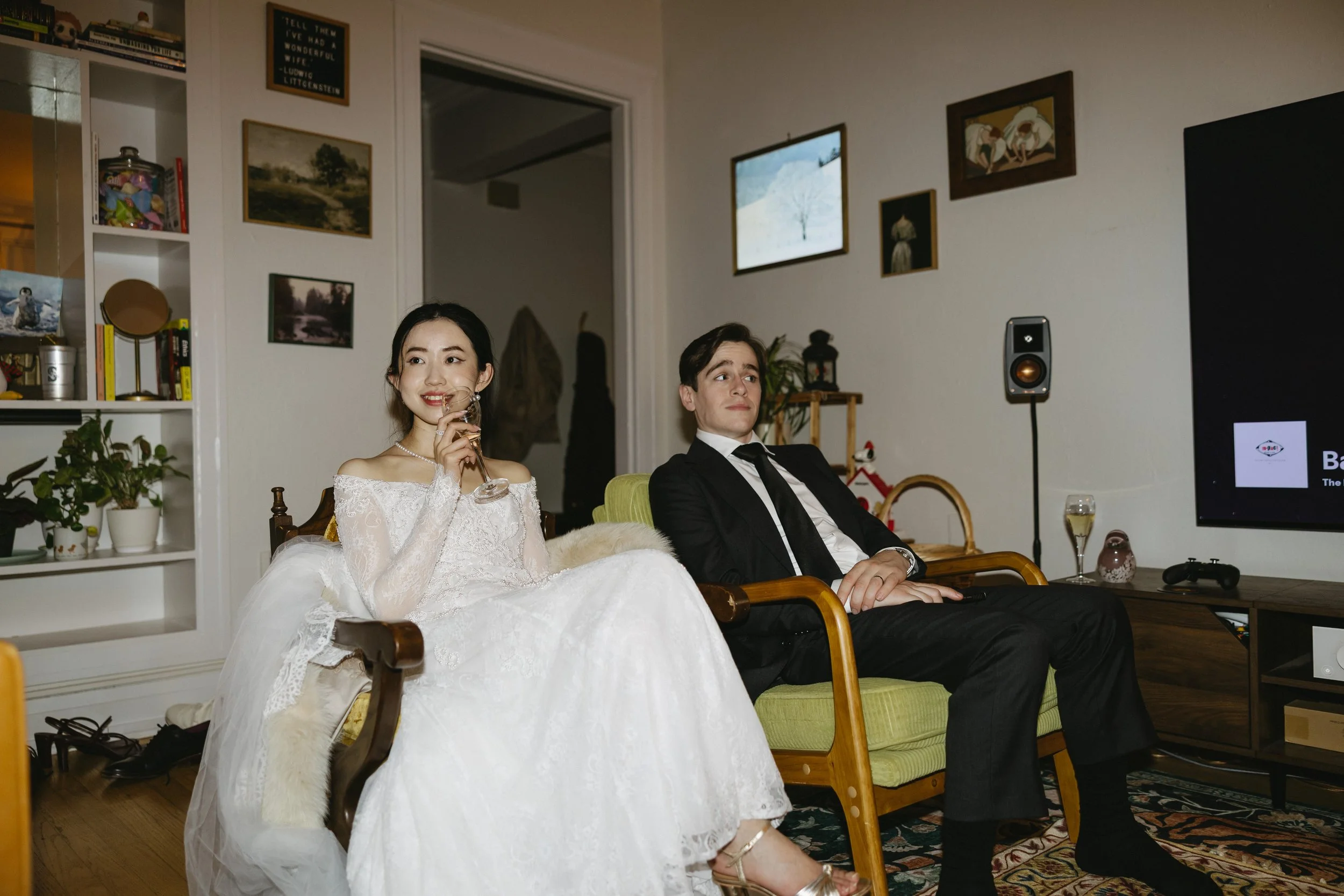 A bride in a white wedding dress and a groom in a black suit sit on chairs in a living room, watching a screen, with a woman holding a glass of wine smiling.