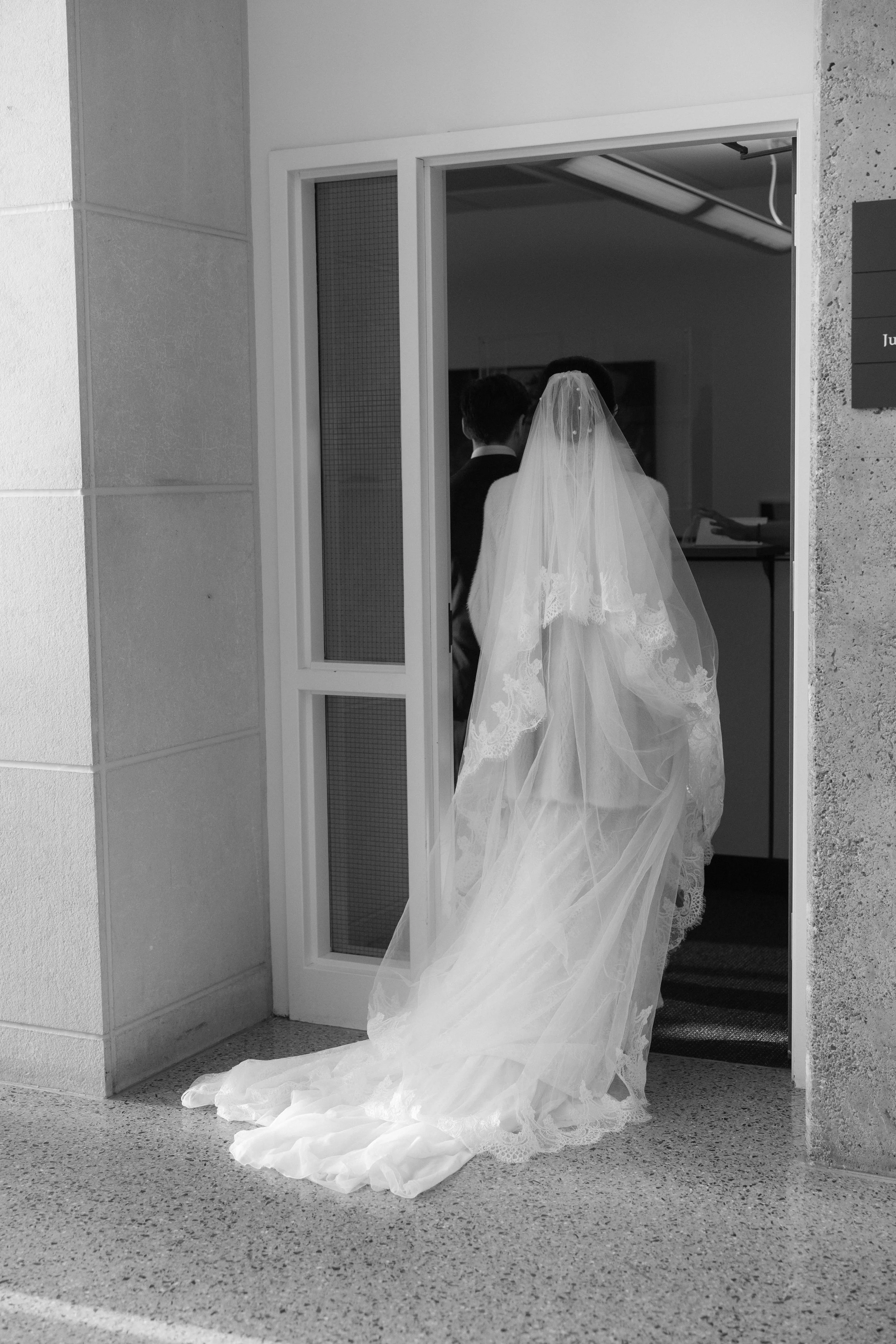 A bride in a wedding dress and veil walking into a building, seen from the back, with two men visible inside.
