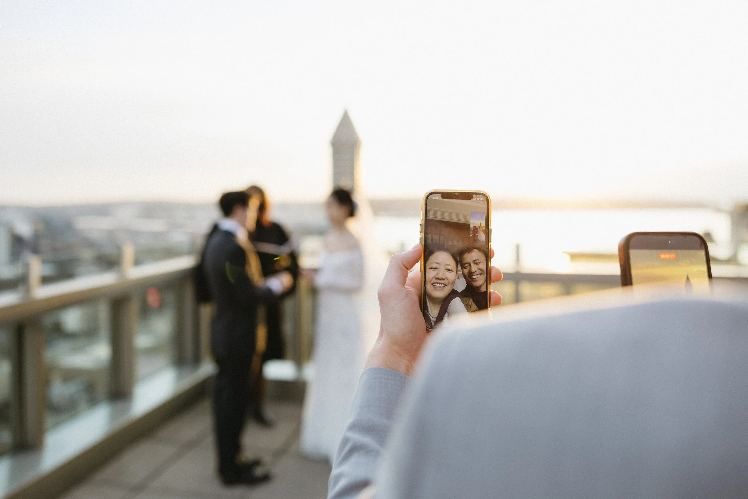 A person taking a selfie of a couple at a wedding on a rooftop with a city skyline in the background, during sunset.