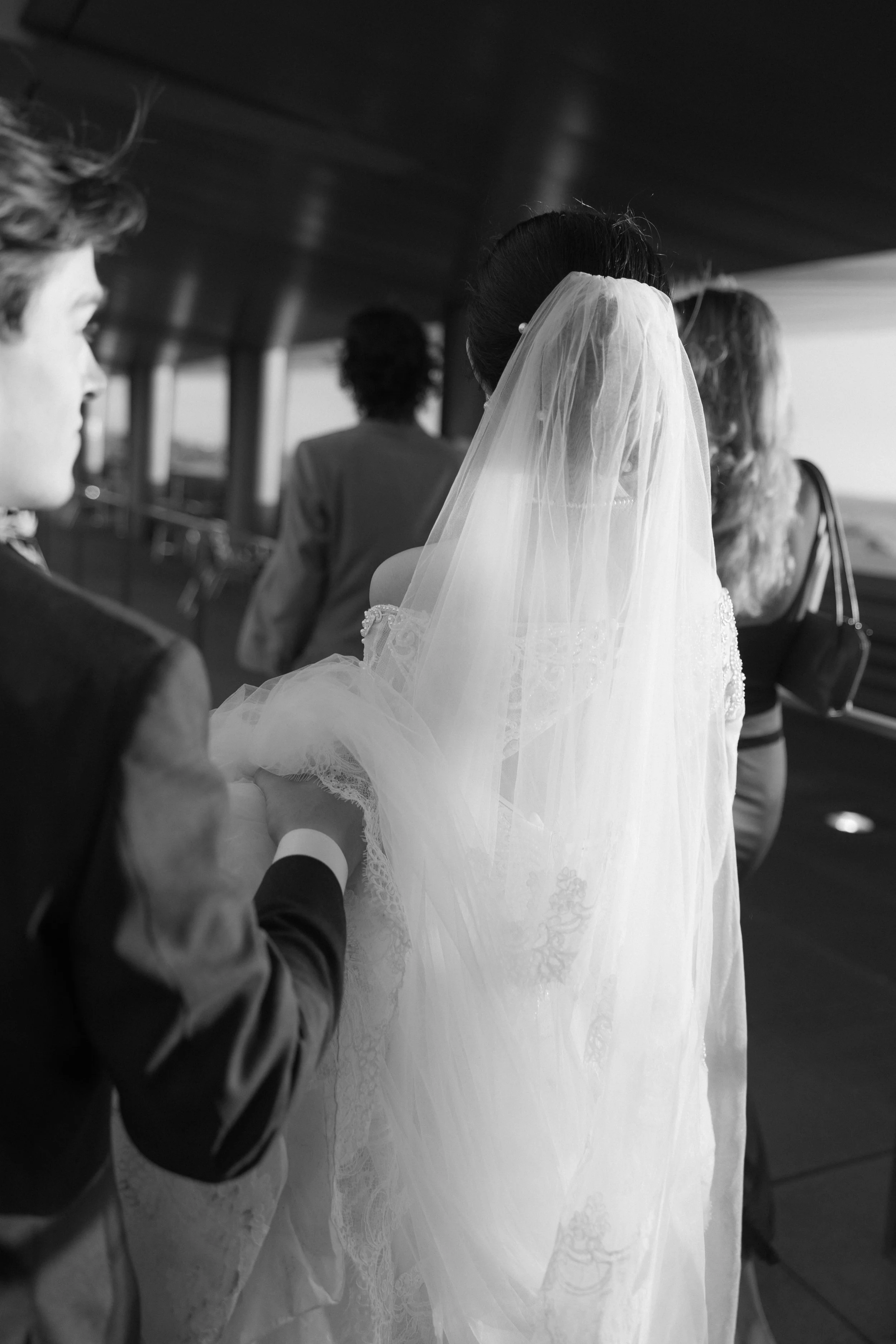 Bride with veil and wedding dress holding hands with a man dressed in formal attire, people in the background, possibly on a boat or outside area.