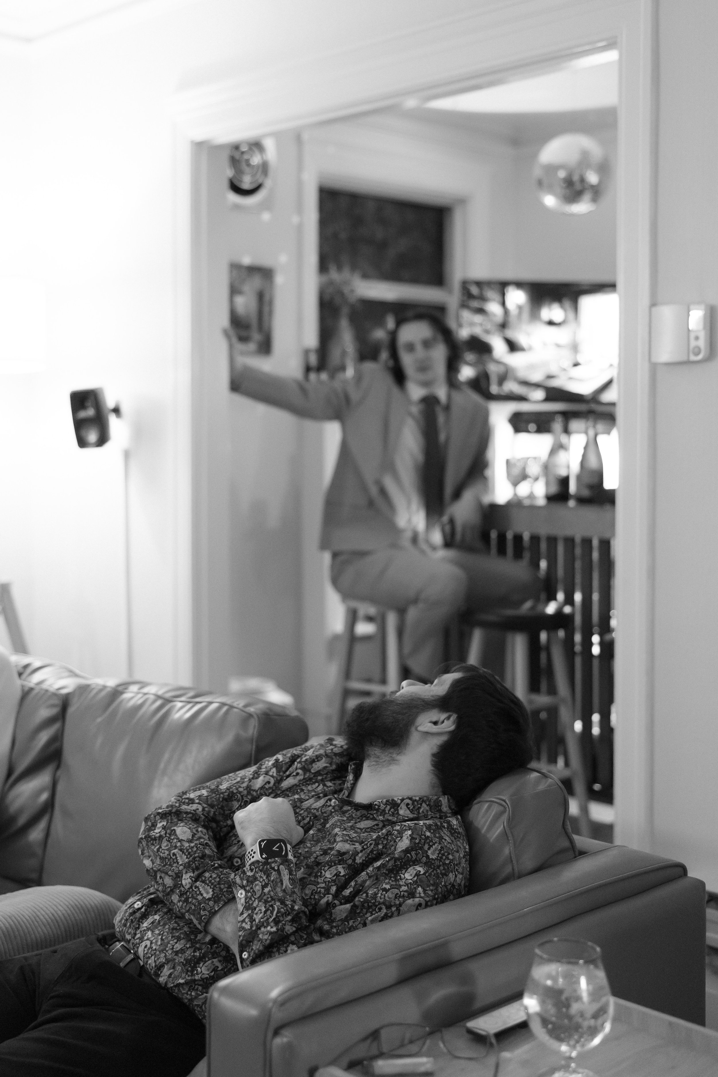 A man with a beard and patterned shirt is sleeping on a sofa, looking relaxed. In the background, a woman with dark hair, wearing a suit and tie, is sitting on a barstool in a kitchen area, gesturing with her right arm. The room appears to be a livin