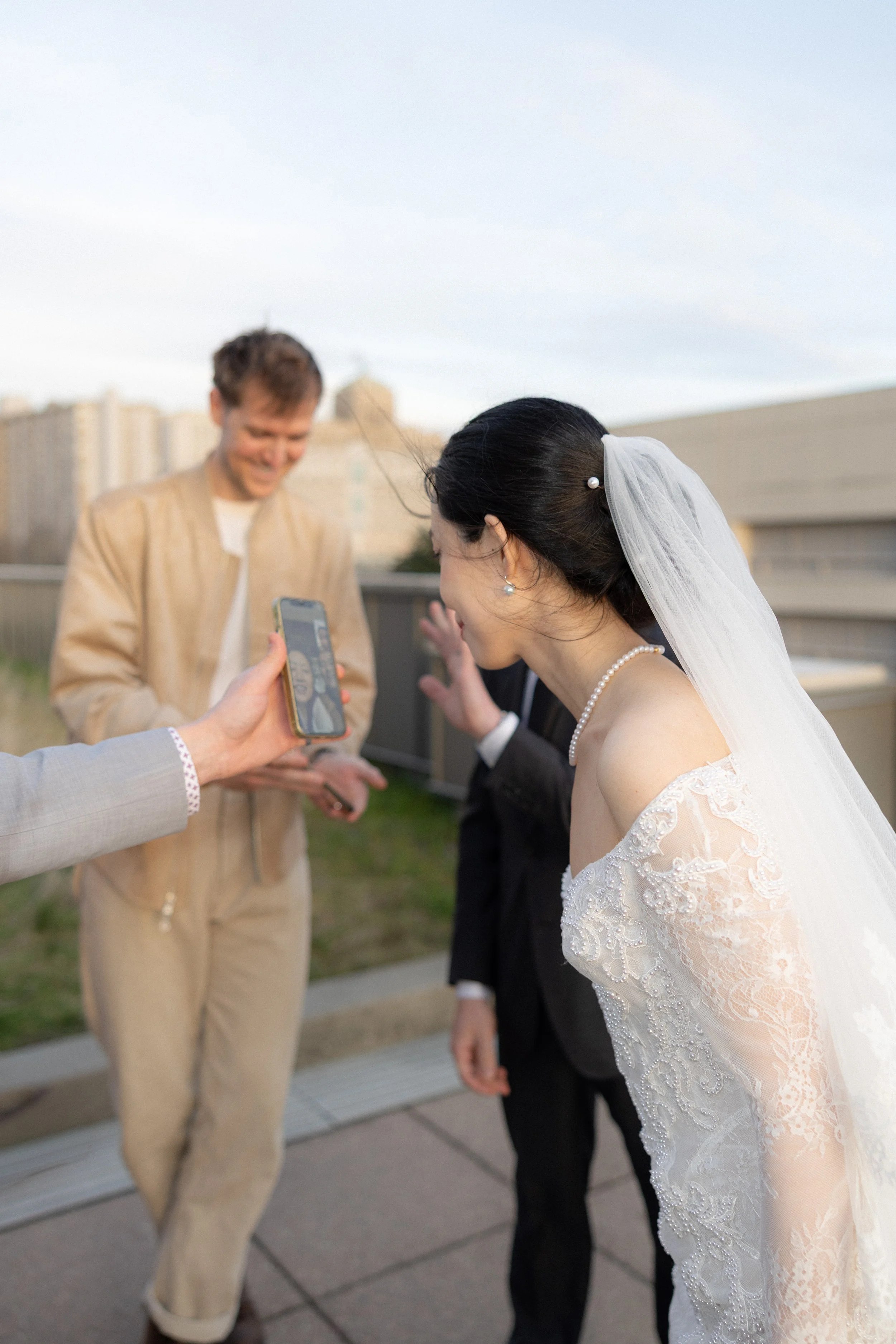 A bride in a white lace wedding dress and veil taking a selfie with a groom dressed in a black suit on a rooftop.