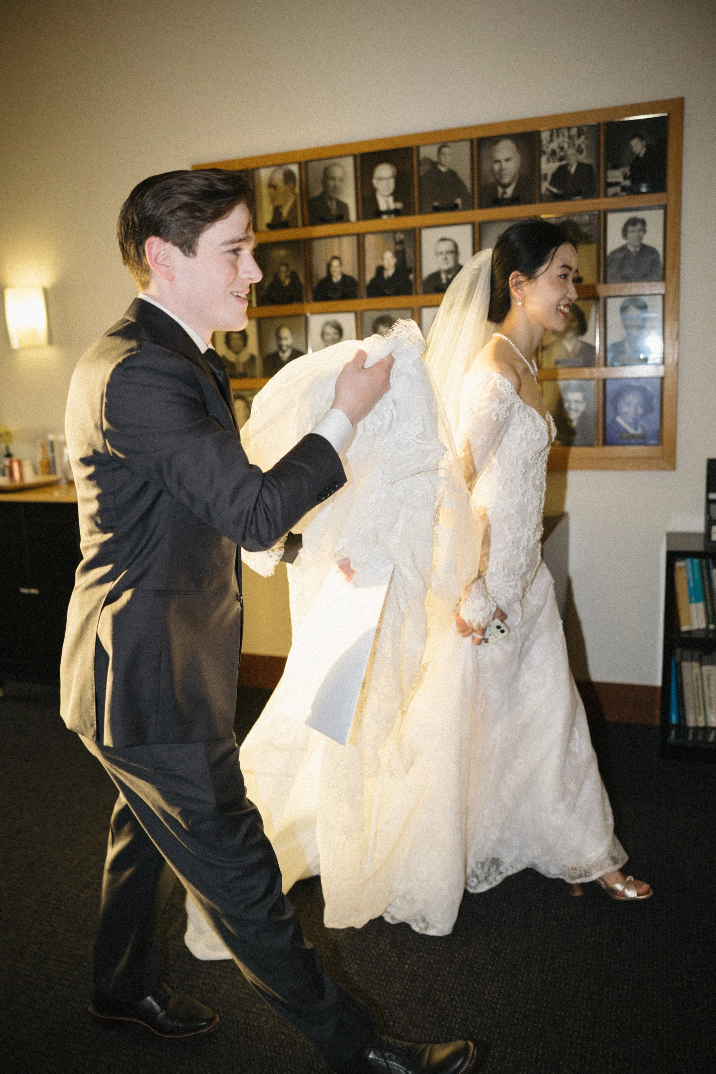 A groom helping a bride with her wedding dress in a room decorated with black and white framed portraits.