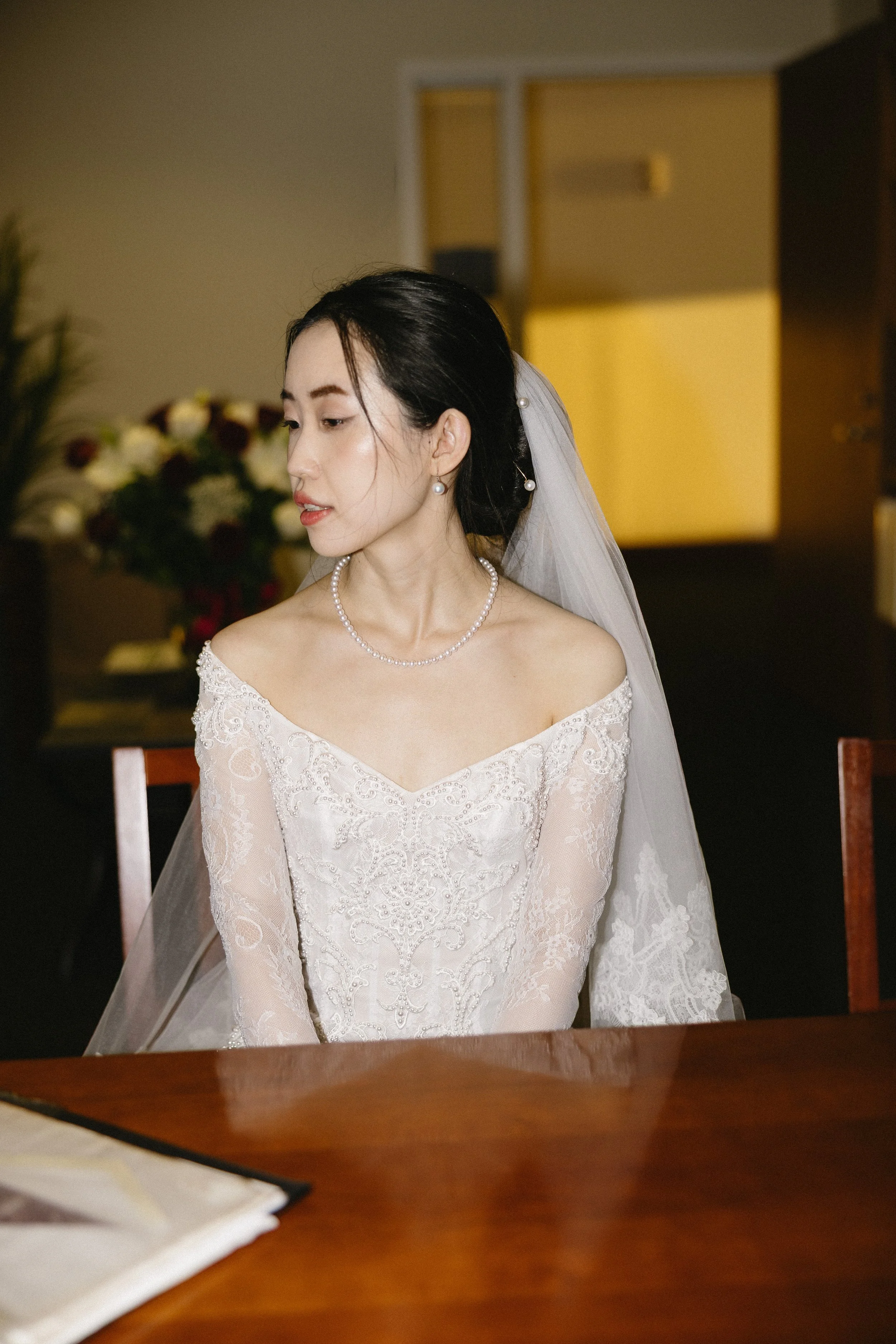 A bride in a wedding dress with off-the-shoulder lace details, wearing pearl jewelry, sitting at a table indoors with a yellow-lit background and a bouquet of flowers in the distance.