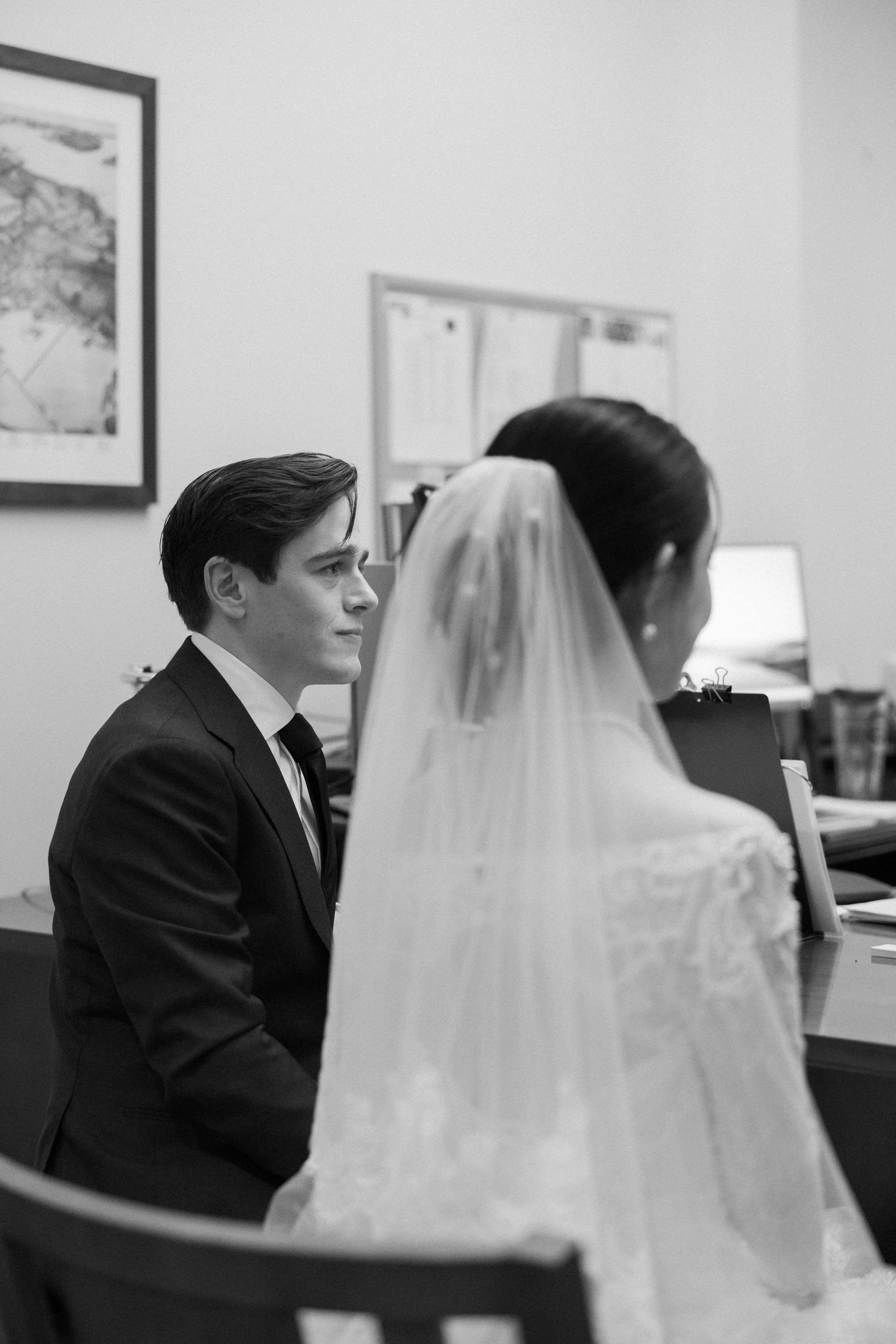 A black and white photo of a man and woman sitting at a desk during a wedding ceremony. The woman is wearing a wedding dress and veil, and the man is in a suit. They are facing each other with focused expressions.