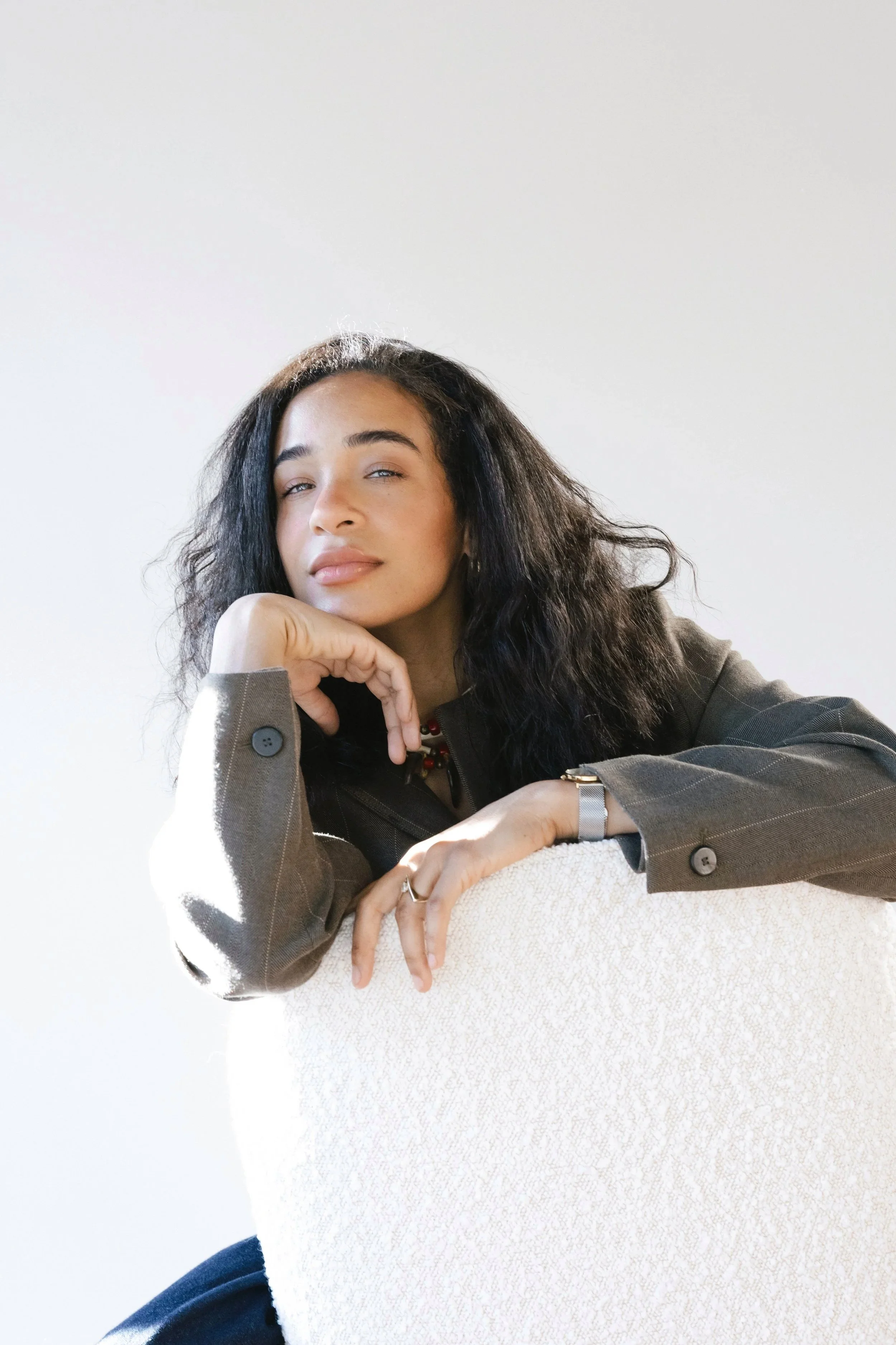A young woman with dark curly hair and light skin is resting her head on her hand, wearing a brown blazer and sitting on a chair against a plain white background.