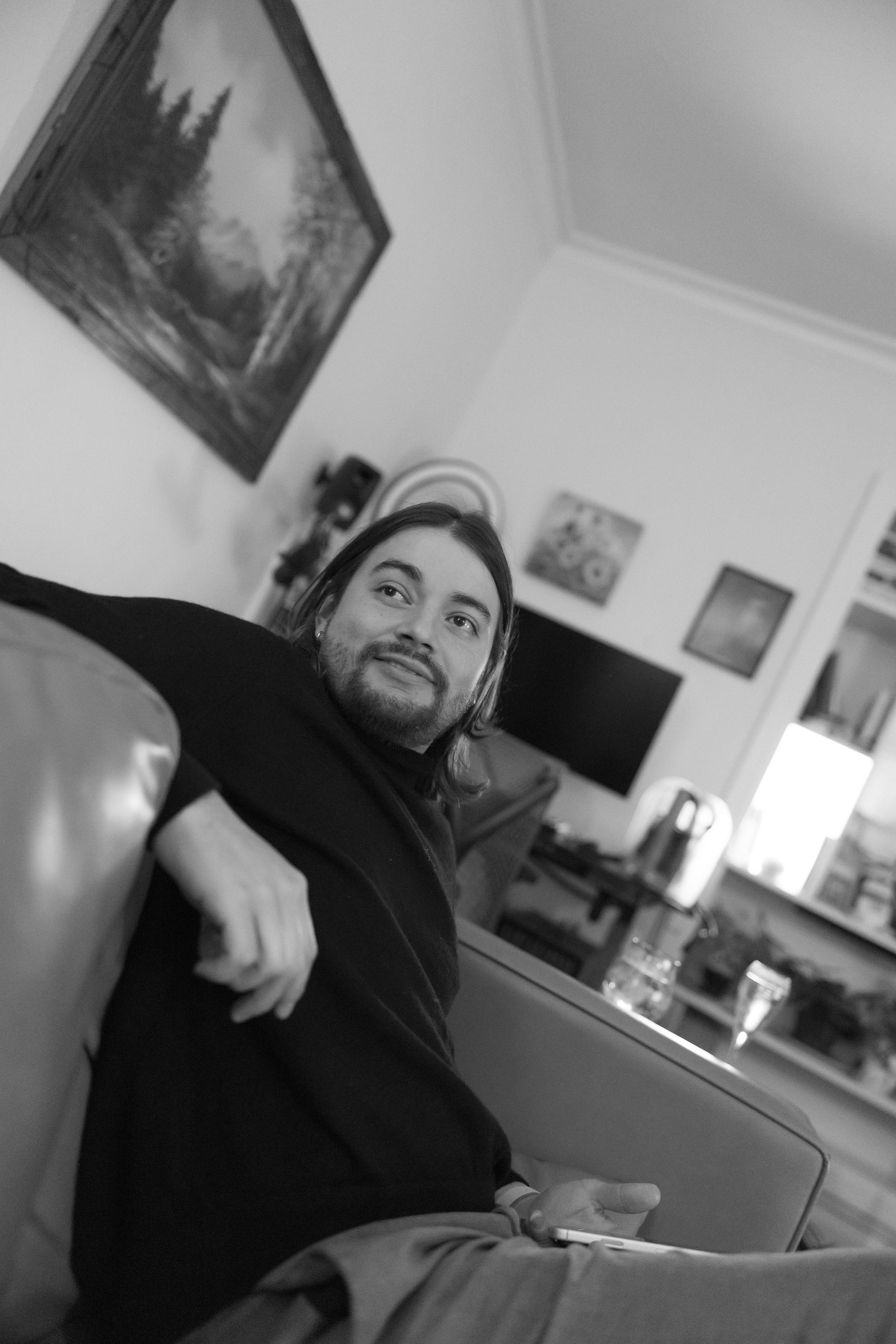 A man with long hair and beard sitting on a couch in a living room, holding a phone in his hand and smiling, with paintings and furniture visible in the background.