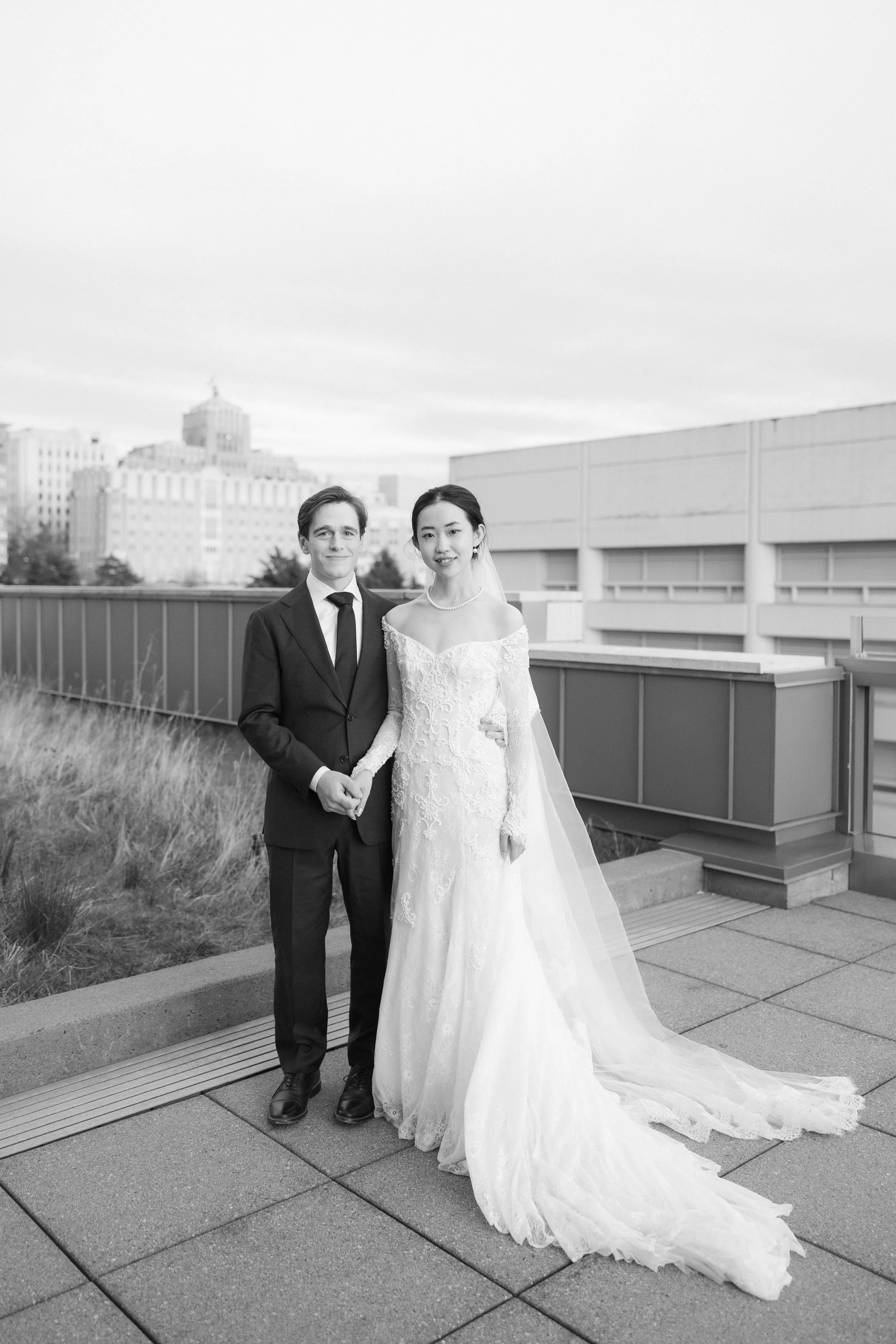 A bride in a lace wedding gown and veiled earrings standing next to a groom in a suit, holding hands on an outdoor rooftop with city buildings in the background.