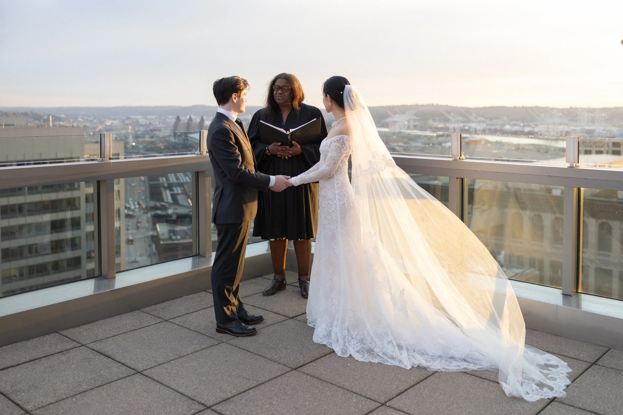 A wedding ceremony with a bride and groom holding hands on a rooftop at sunset, officiated by a woman with an open book.