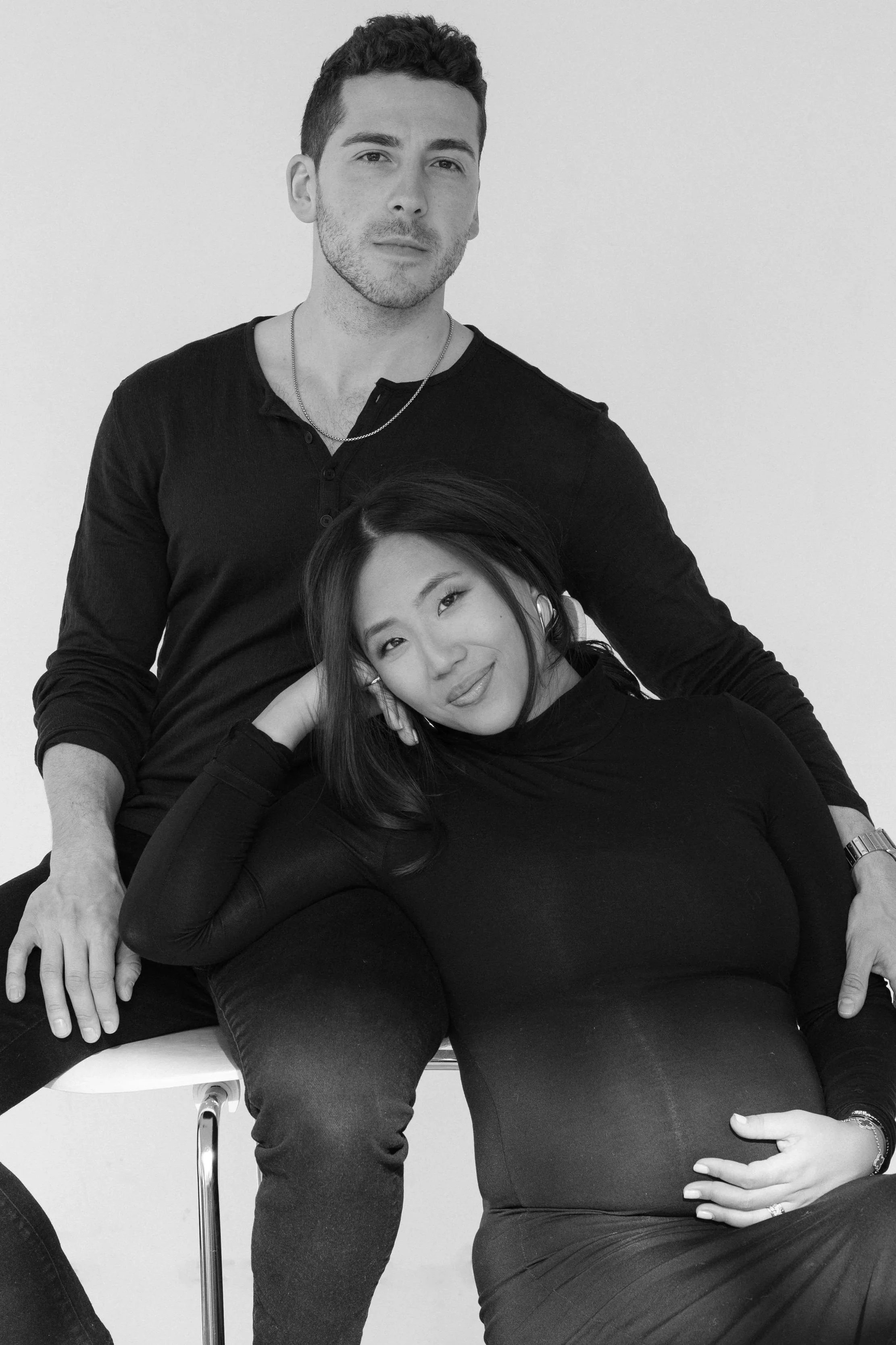 A black and white photo of a man and a pregnant woman posing together. The man is standing behind the woman, with one hand on his knee and the other on the seat. The woman is seated and resting her head on her hand, with her other hand on her pregnant belly.
