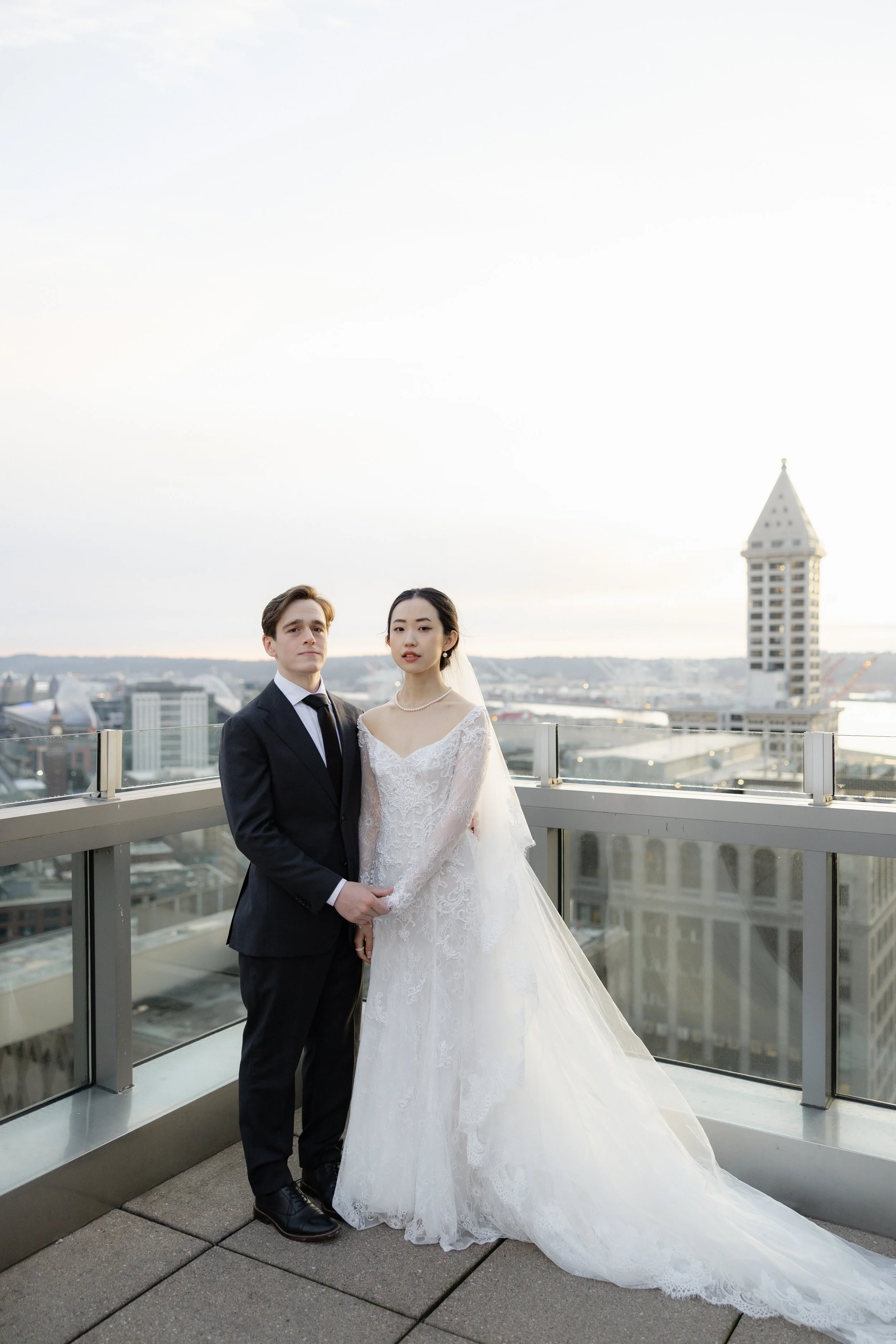 A wedding couple stands on a rooftop with city buildings and a tower in the background, the bride in a white lace wedding gown and the groom in a black tuxedo.
