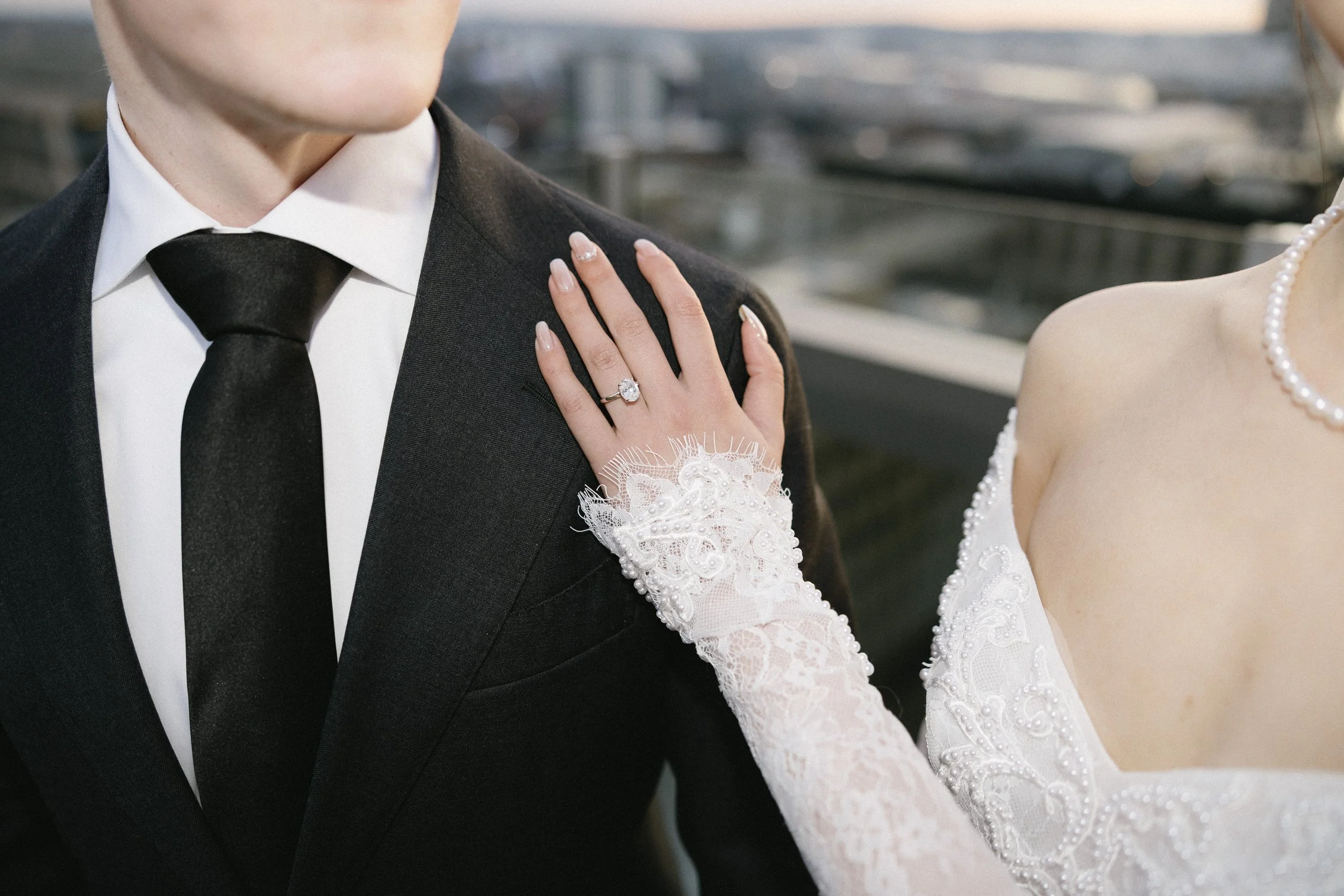 Close-up of a bride and groom on their wedding day, focusing on the bride's hand resting on the groom's shoulder, showing her engagement ring and wedding band. The bride is wearing a lace gown and pearl necklace, while the groom is in a black suit wi