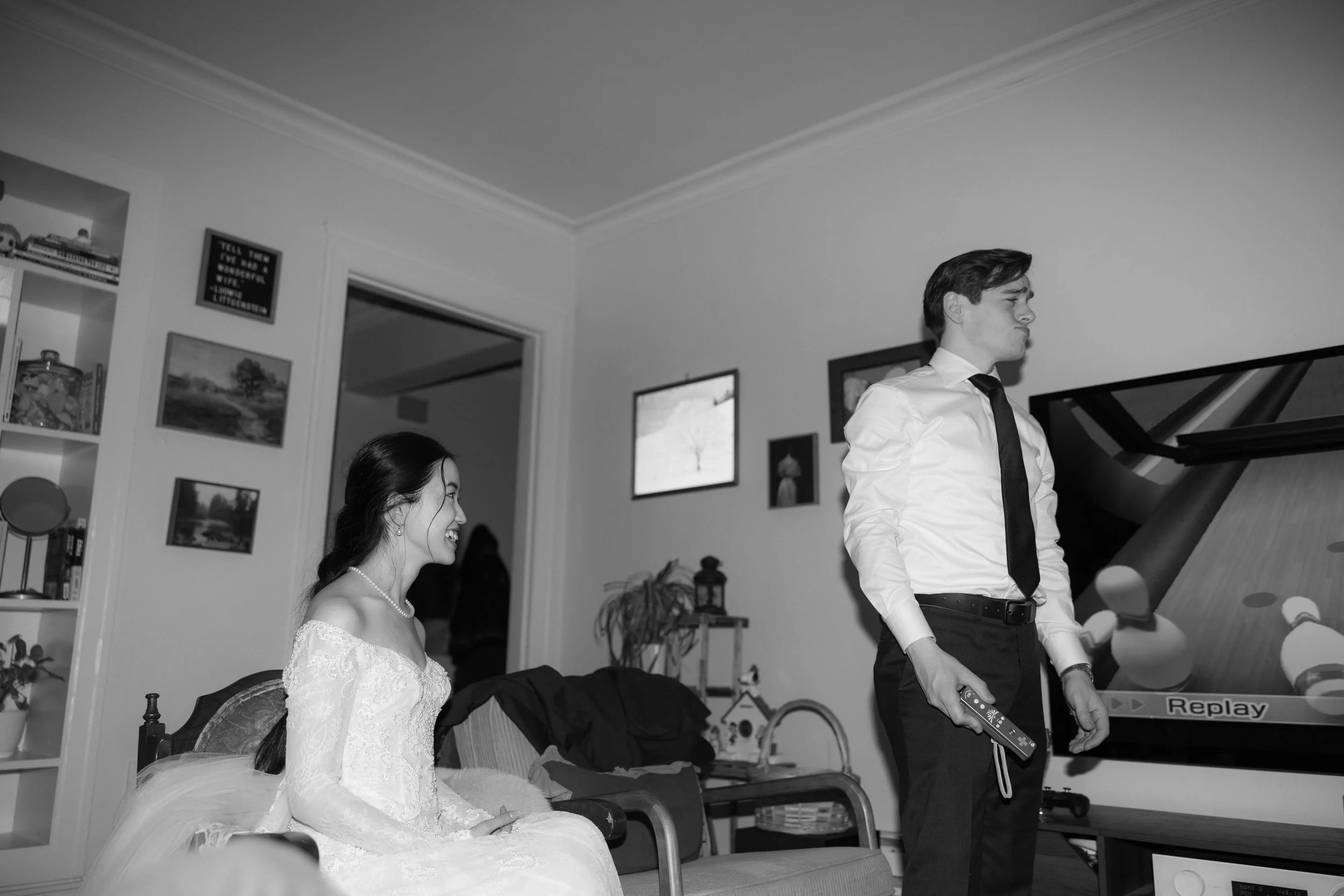 A woman in a wedding dress sitting on a couch, smiling at a man in formal attire playing a video game in a living room.