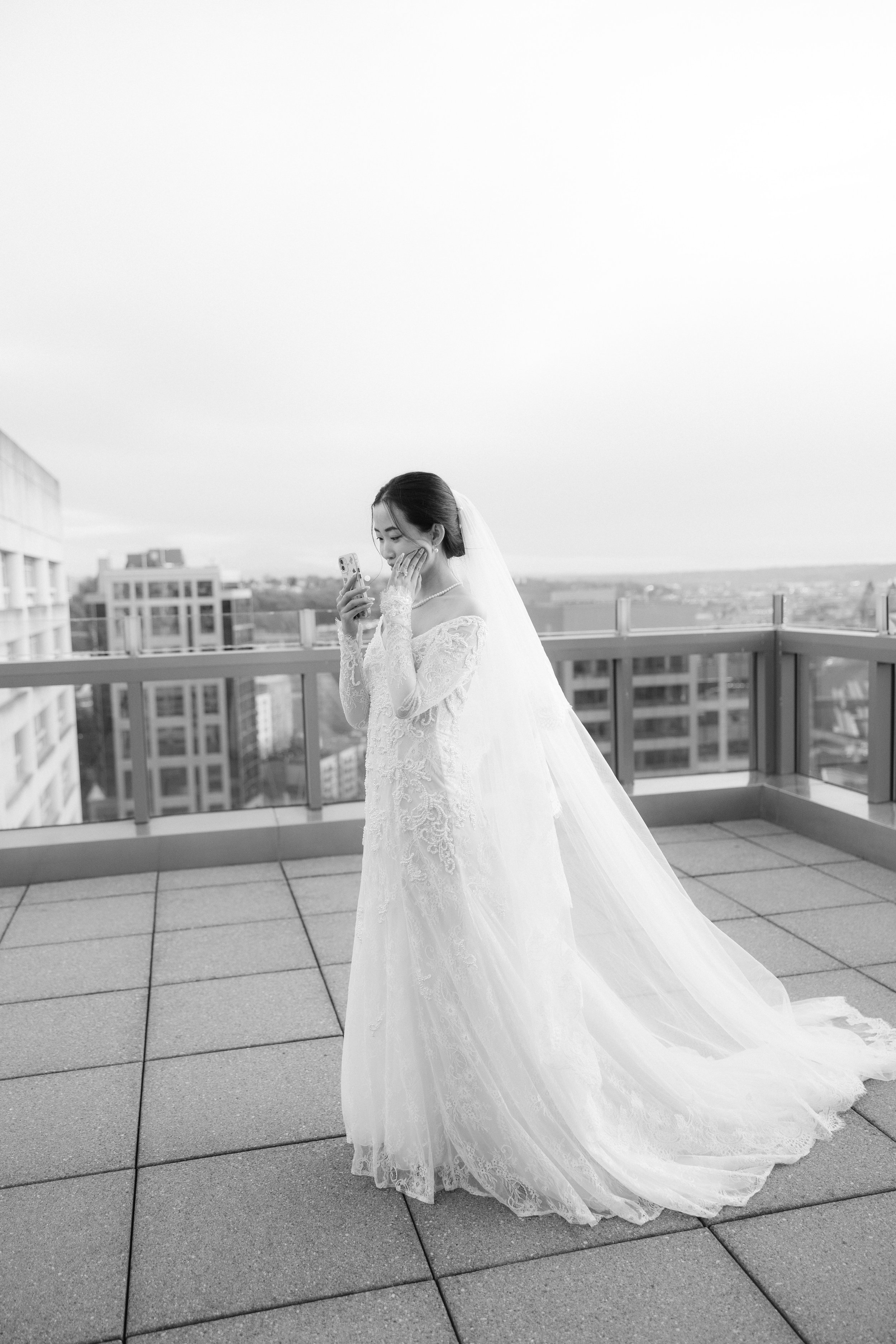 A bride in a wedding dress and veil looking at her phone on a rooftop terrace with city buildings in the background.