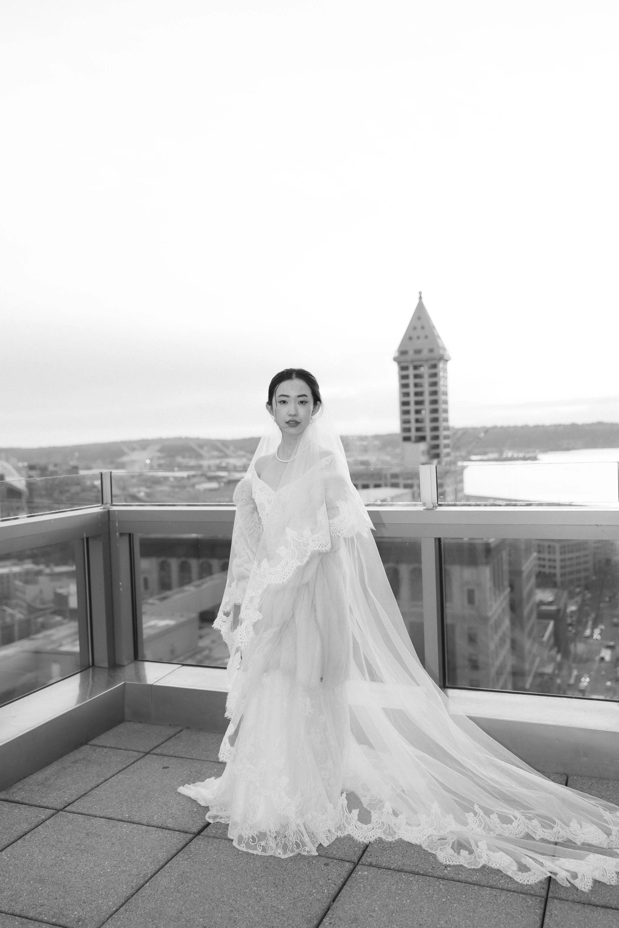 A woman in a wedding dress with a long train posing on a rooftop with city buildings and a tower in the background