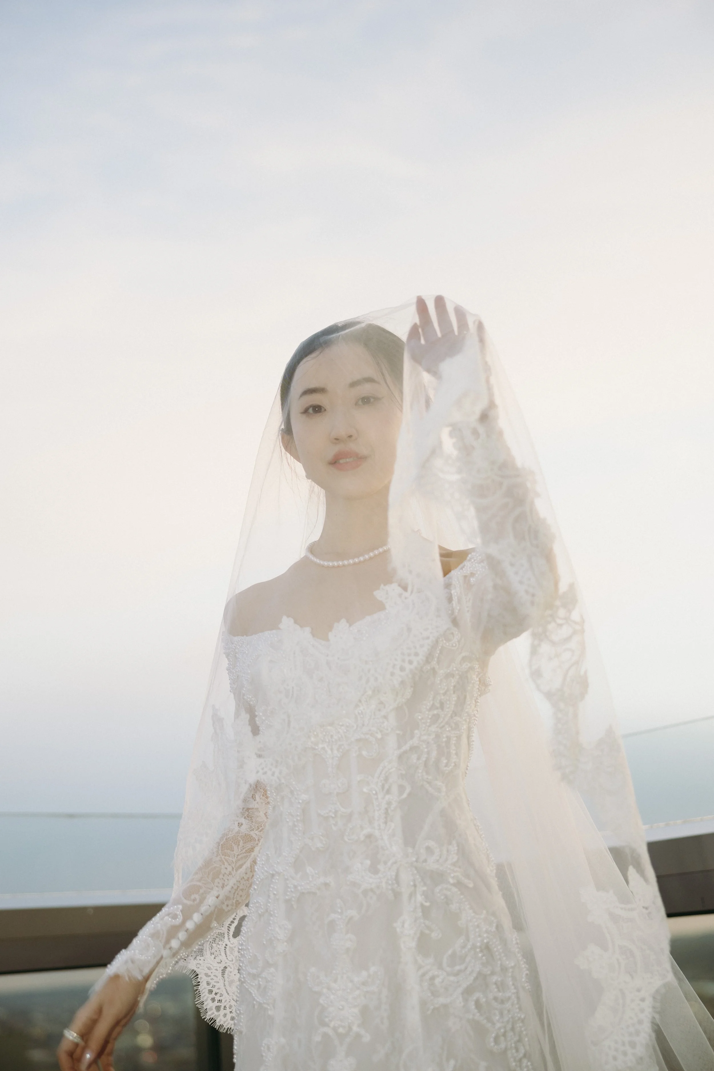 A woman dressed in a wedding gown and veil, standing outdoors against a light sky, holding her veil with her hand.