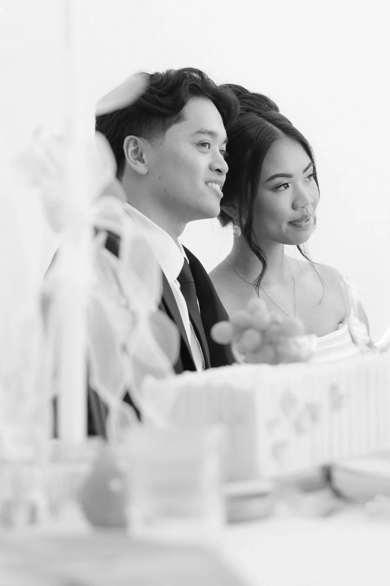 Black and white photo of a happy couple sitting at a table, possibly during a celebration or formal event, with blurred objects and decorations in the foreground.