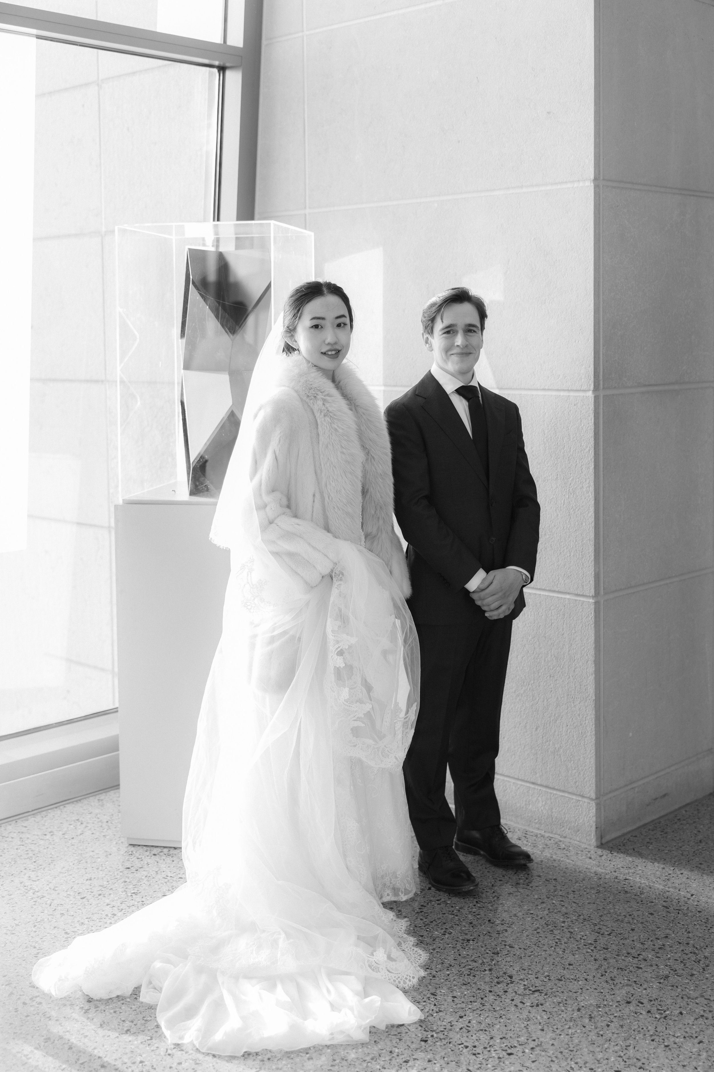 A bride in a wedding dress and a groom in a suit standing together indoors near a window with an abstract art sculpture in the background.