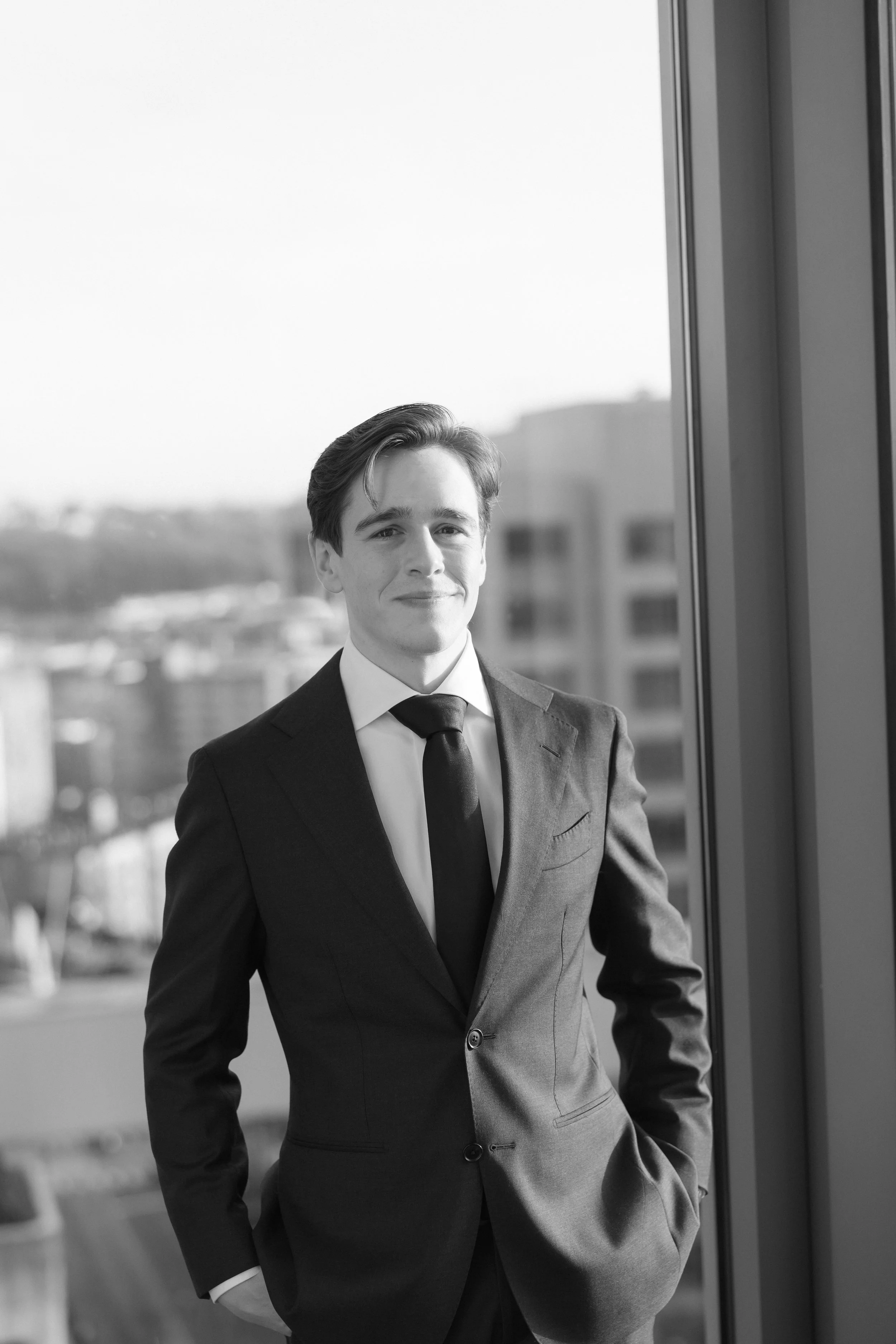 A young man in a suit with his hands in his pockets standing next to a large window, smiling at the camera, with an urban cityscape in the background.