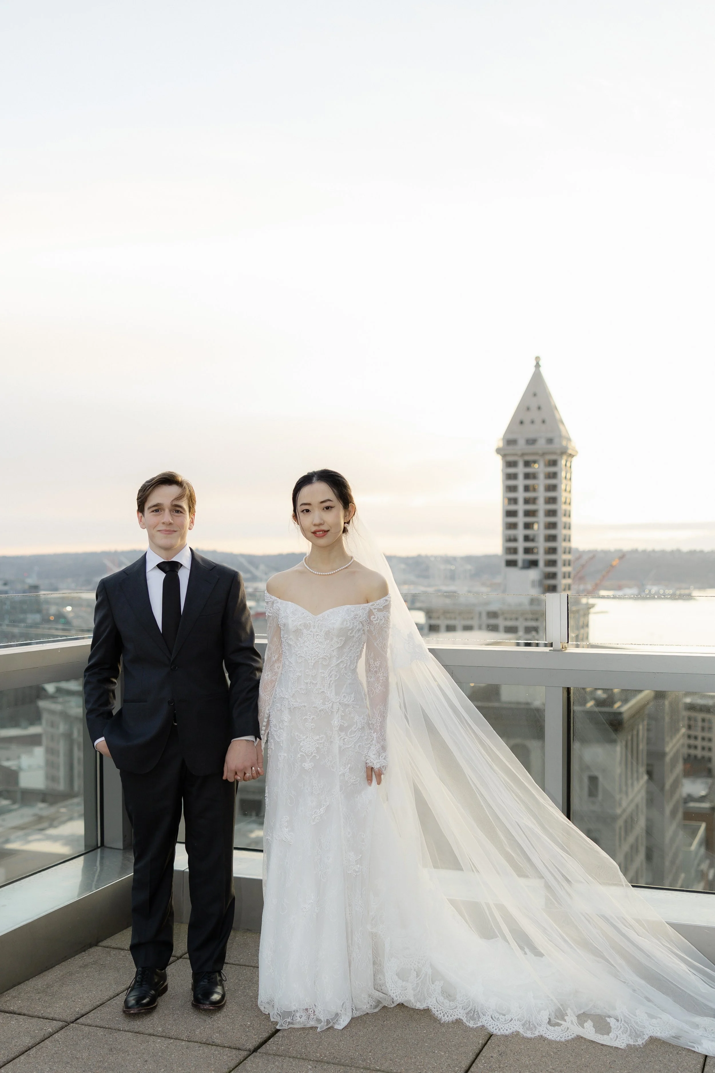 A bride and groom holding hands on a rooftop with city buildings in the background during sunset.