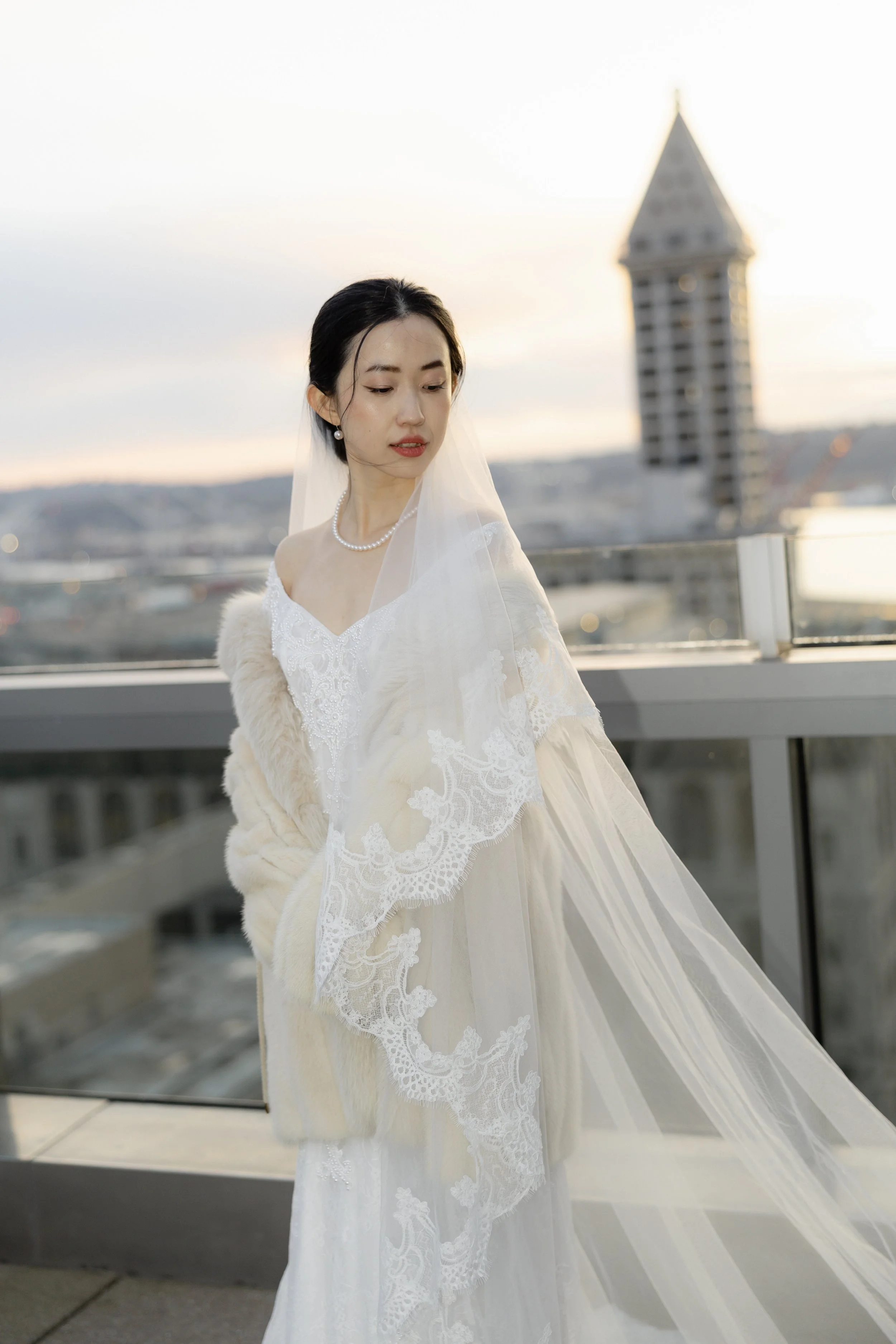 A woman in a white wedding dress with lace details, a pearl necklace, and a lace veil, standing on a rooftop with a cityscape and tall building in the background during sunset.