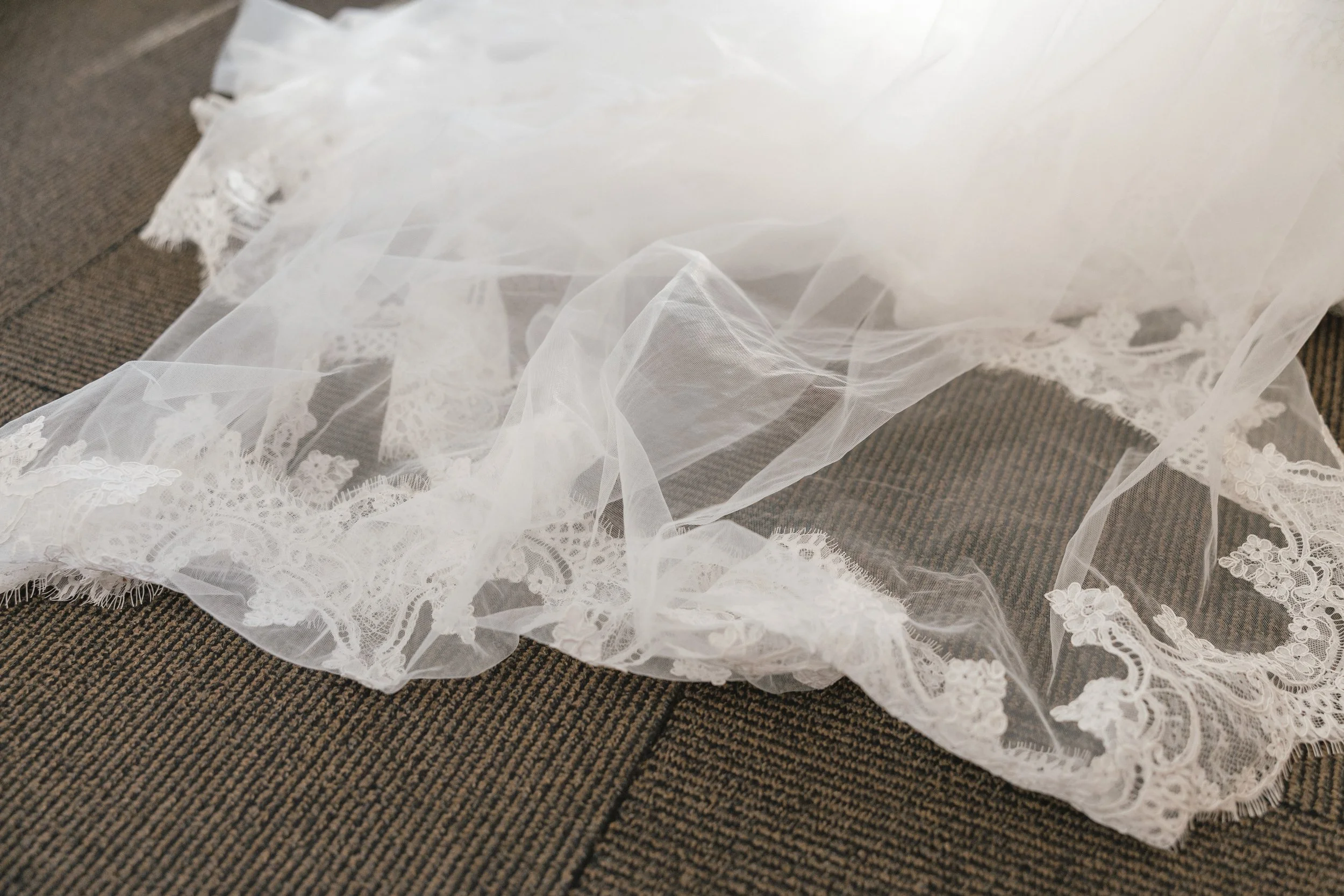 A white, sheer lace and tulle bridal veil with intricate embroidery, laid out on a brown textured carpet.