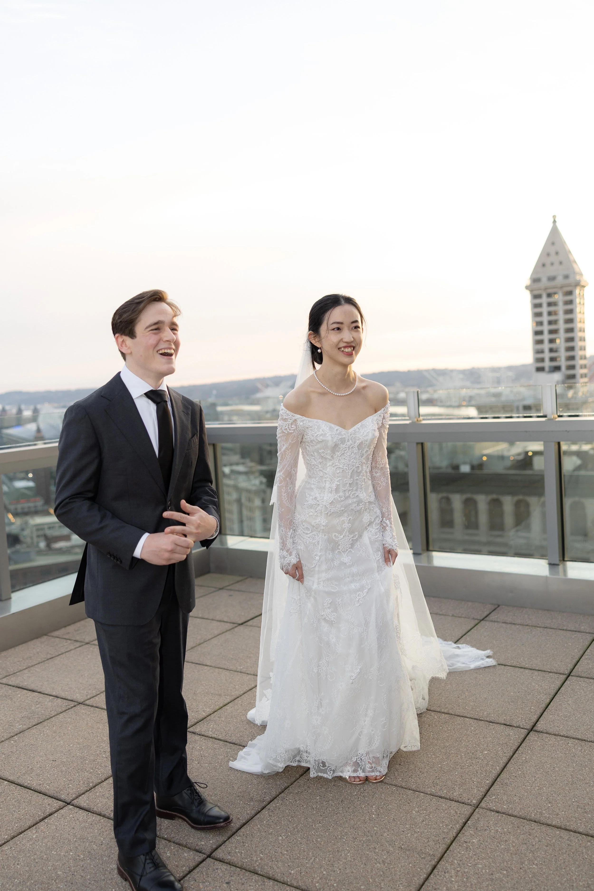 A bride in a white wedding dress and a groom in a black suit on a balcony with a city skyline in the background during sunset.