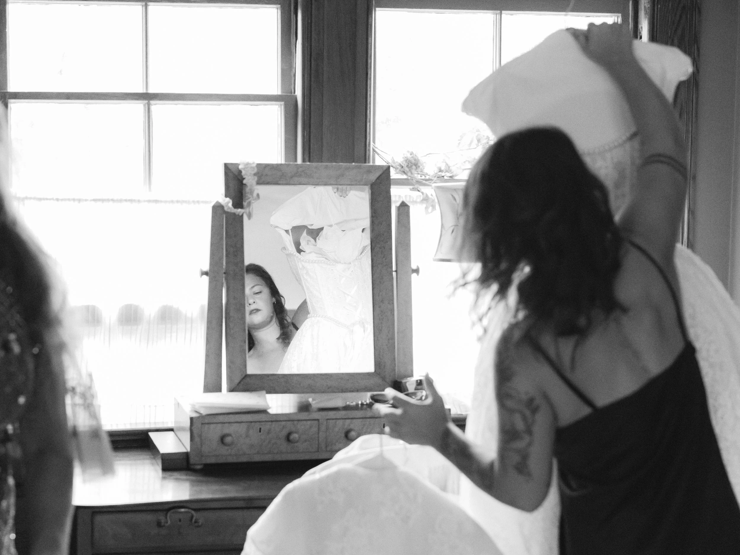 A woman with dark hair looking at her reflection in a mirror in a room with large windows, with a person holding a white umbrella nearby.
