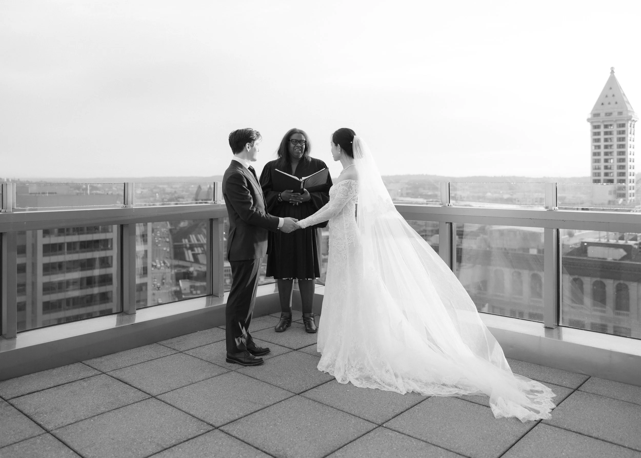 A black-and-white photograph of a wedding ceremony on a rooftop, showing a bride and groom holding hands and facing each other, with an officiant in the middle, against a city skyline background.