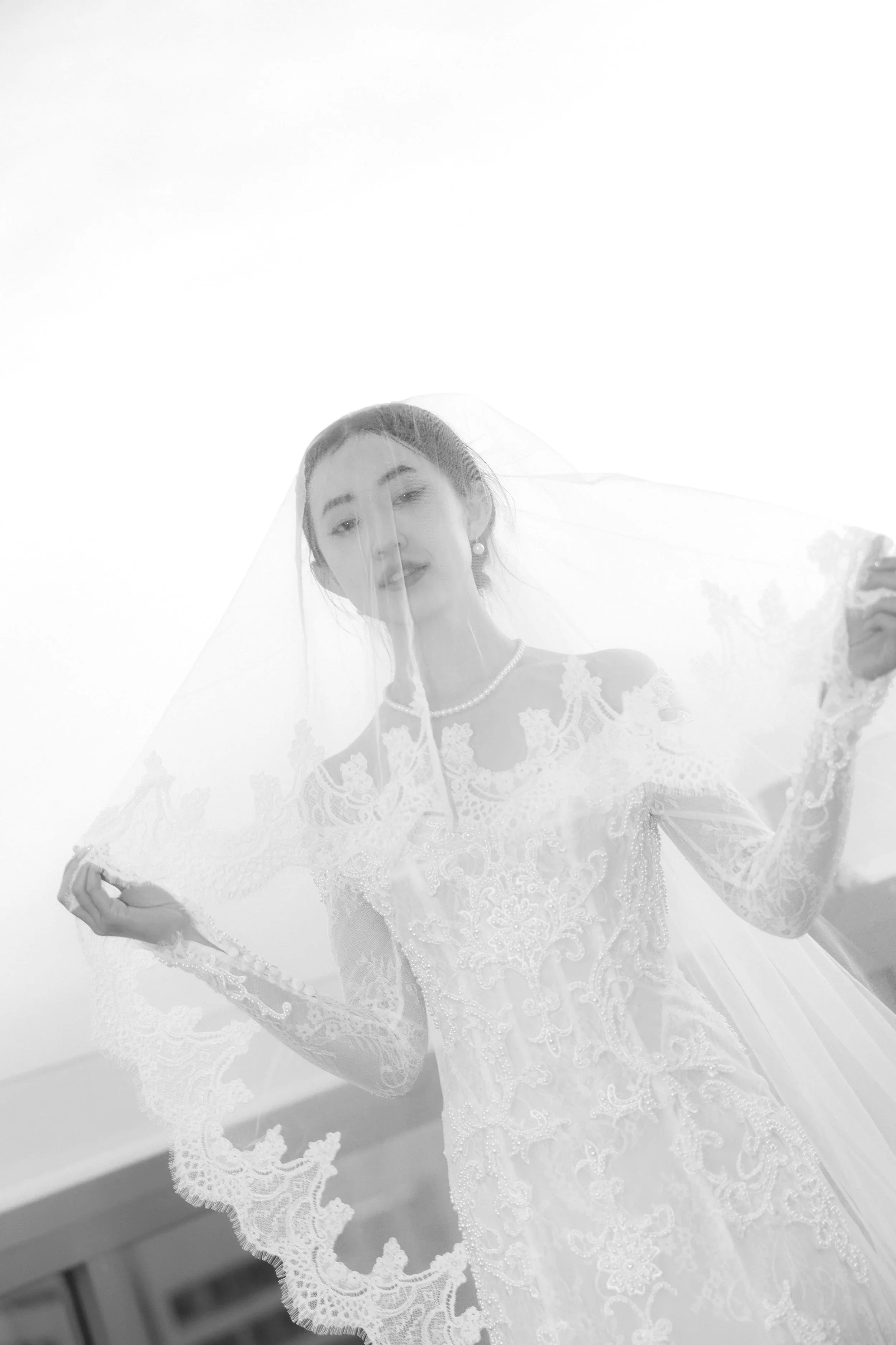 A woman in a lace wedding dress holding a veil in an outdoor setting. Editorial wedding photography.