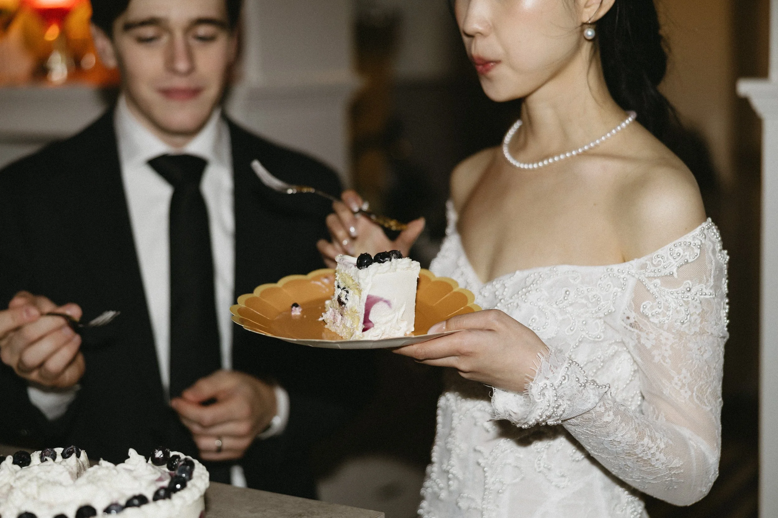 Bride in a white lace dress and pearl jewelry holding a slice of cake on a gold plate, while man in a black suit and tie stands nearby, at a wedding celebration.