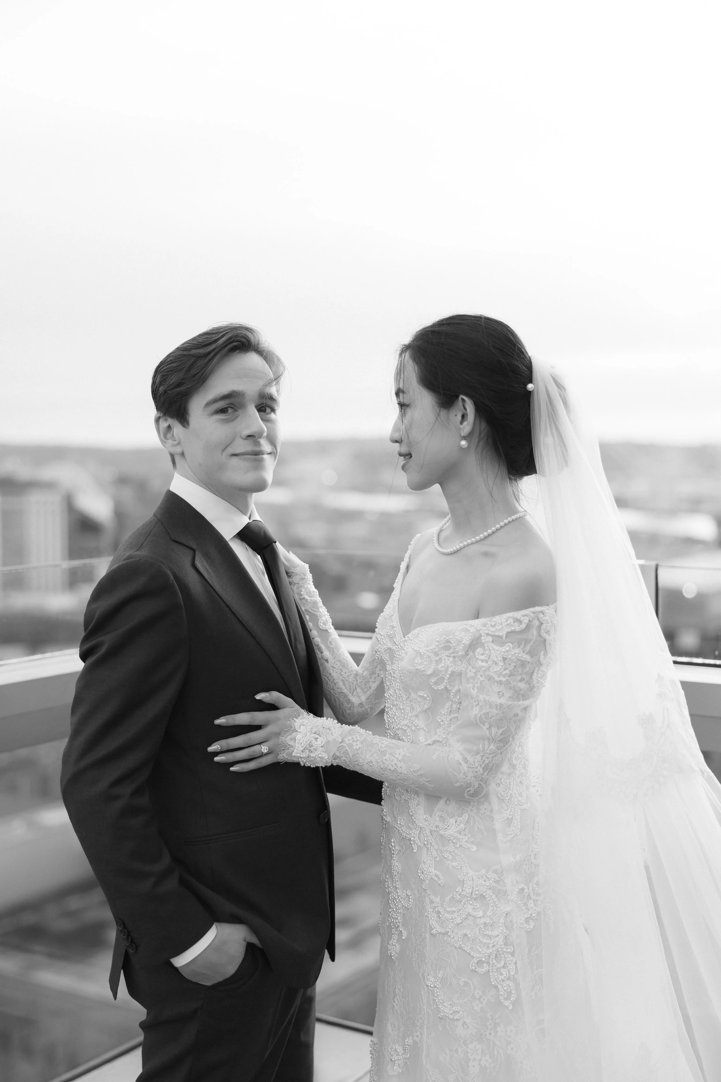 Black and white photo of a bride and groom on a rooftop, facing each other. The groom wears a suit and tie, and the bride wears a lace wedding gown with a veil. The bride has her hand on the groom's chest, and they are looking at each other.