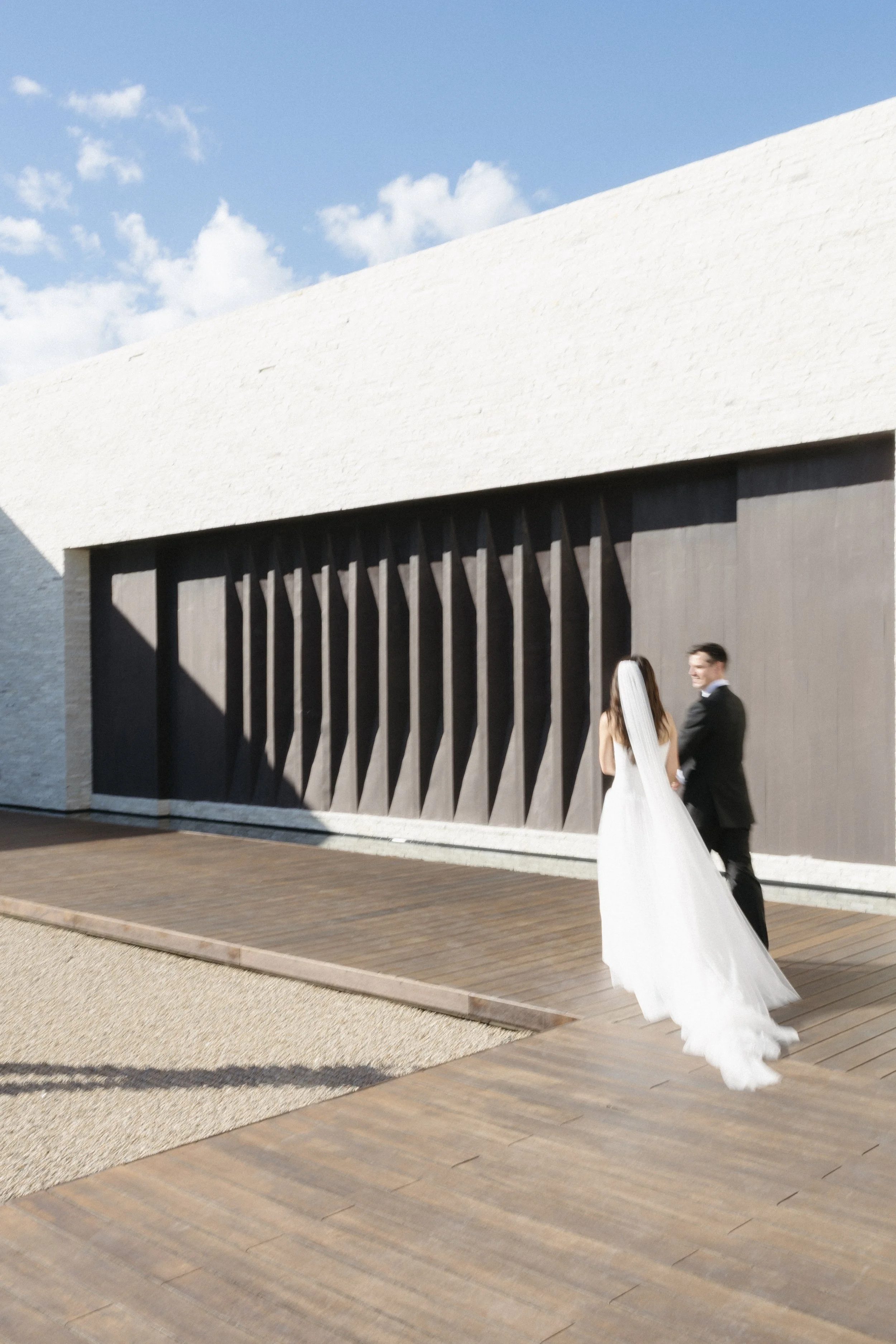 A bride and groom walking hand in hand outside a modern building with a white roof and dark wooden door, on a sunny day.