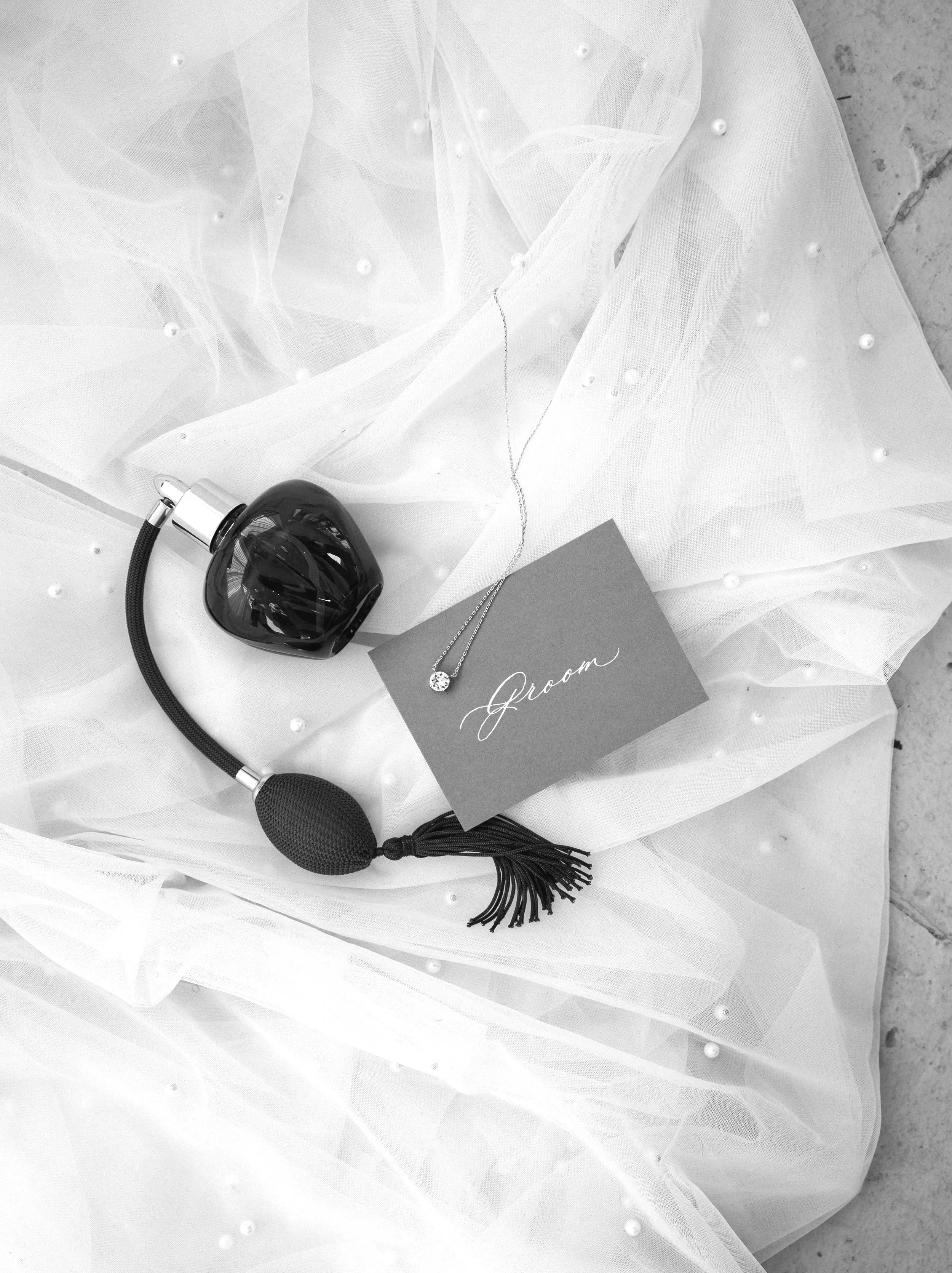 A black perfume bottle, a black textured massage tool, a necklace with a gemstone, and a card with 'Groom' written in cursive, all placed on sheer white fabric with small pearls scattered on it.
