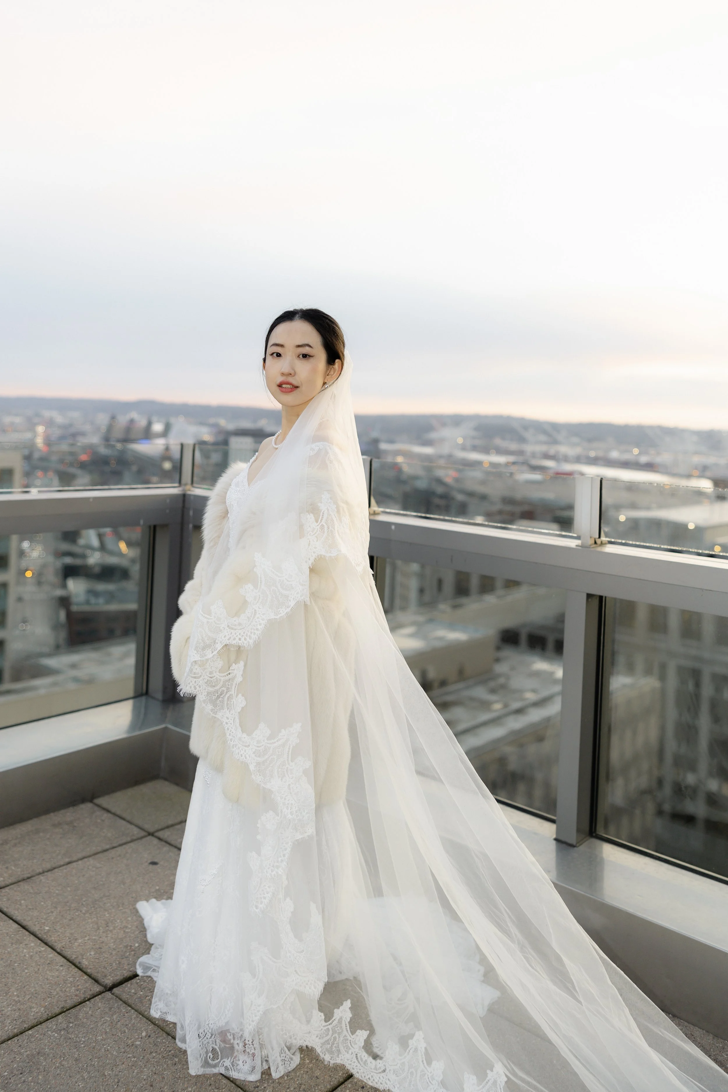 A woman in a white wedding dress and veil standing on a rooftop with a city view in the background during sunset.