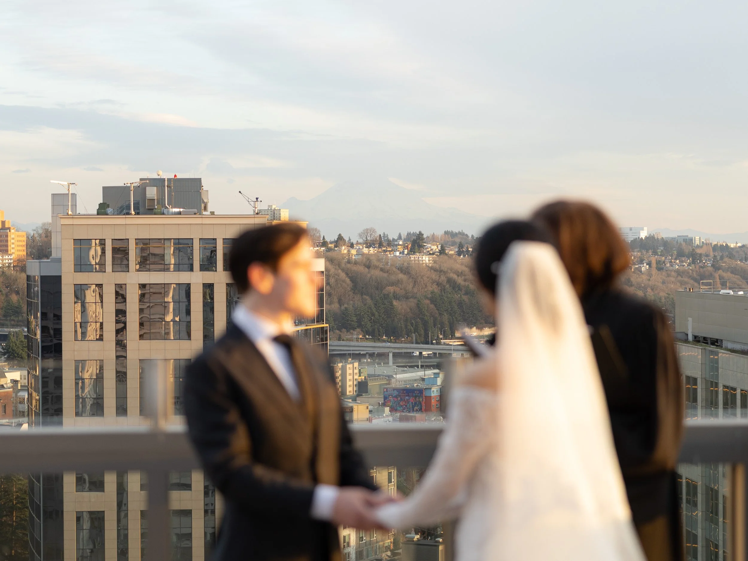 A wedding ceremony taking place on a rooftop terrace with cityscape and hills in the background, featuring a bride in a white dress and veil holding hands with the groom in a black suit, both facing each other, along with a woman on their right.