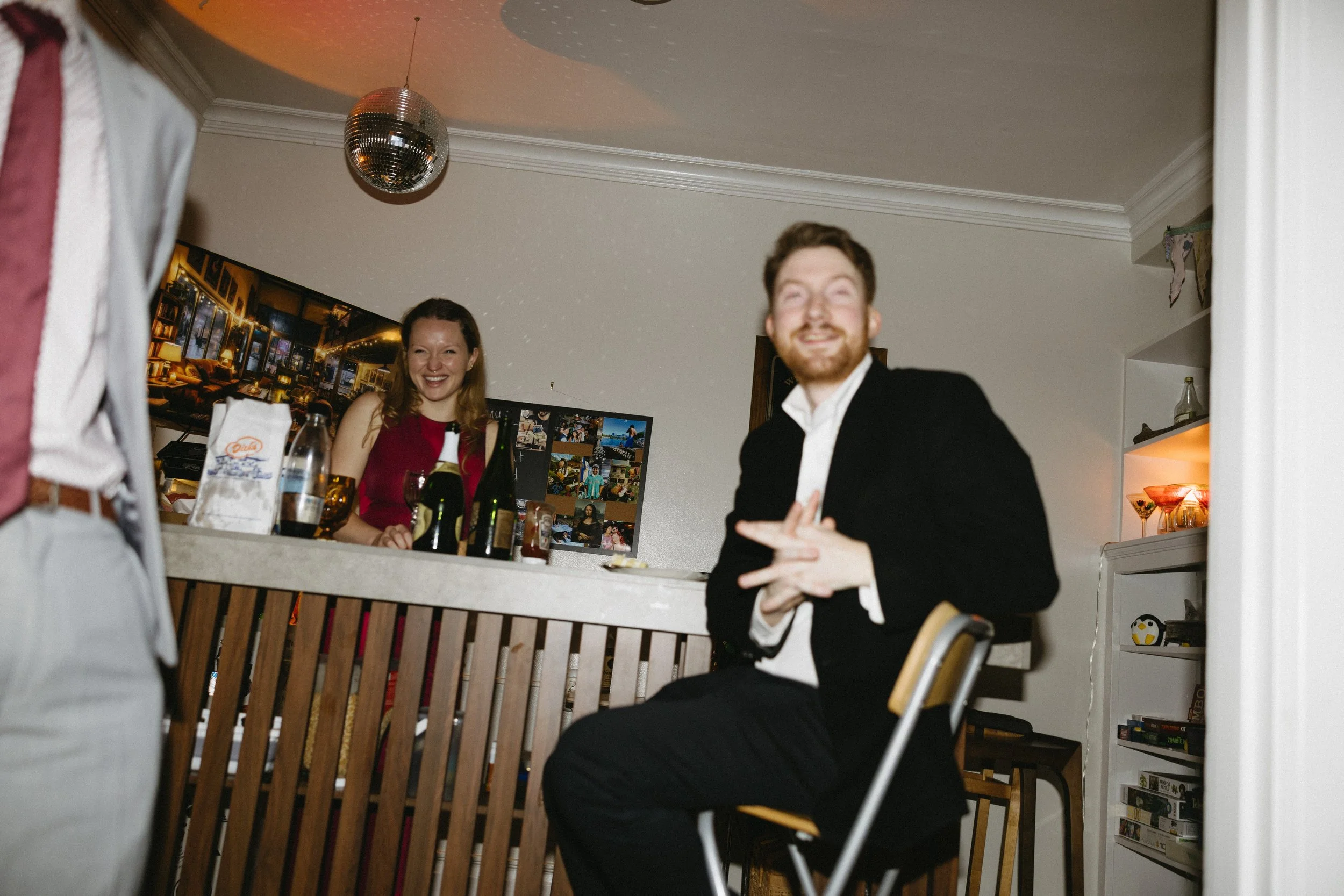 People at a party in a living room, smiling and enjoying drinks. One woman is standing behind a counter with bottles, and a man dressed in a tuxedo is sitting in the foreground.