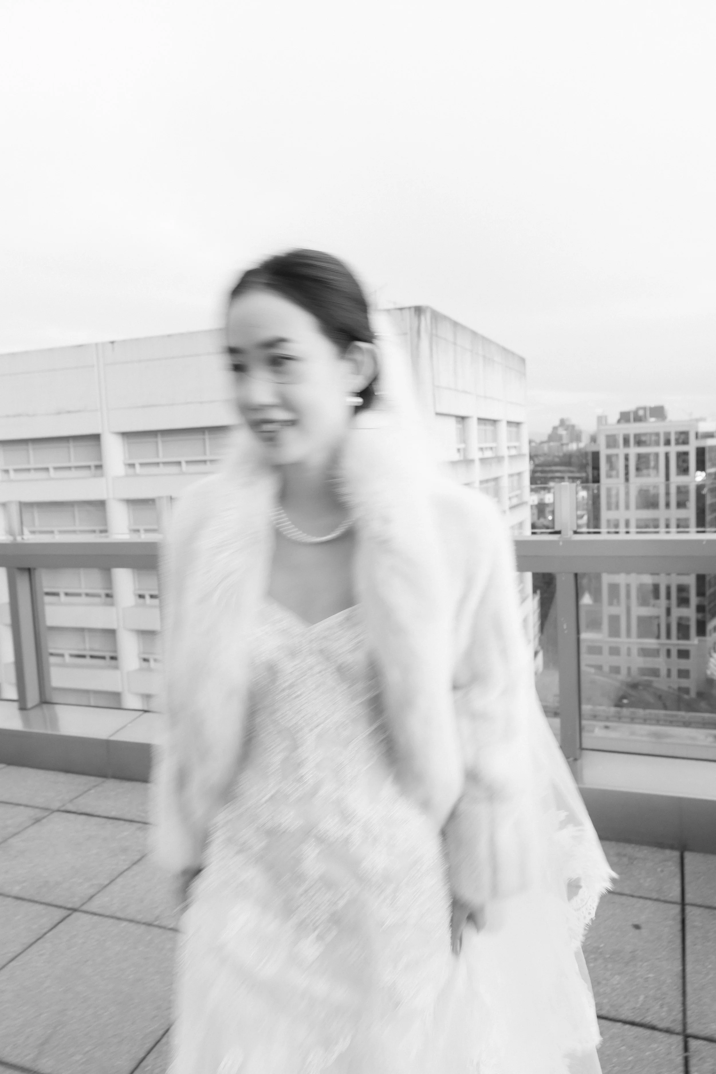 Blurry black and white photo of a woman wearing a fur coat and a dress with a string of pearls, standing on a rooftop with buildings in the background.