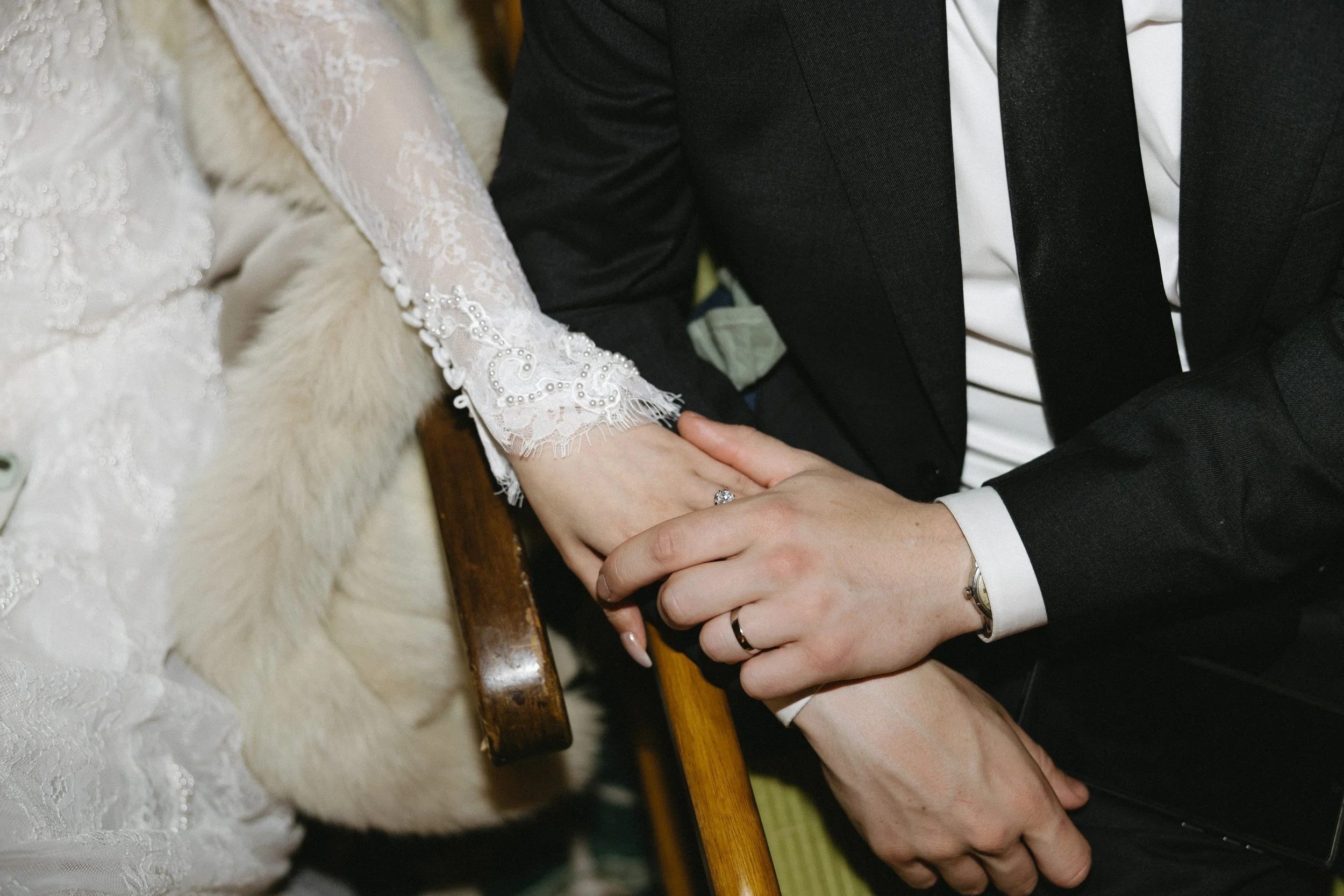 A bride and groom holding hands, with the bride wearing a lace wedding dress and the groom in a black tuxedo, sitting on a wooden bench.