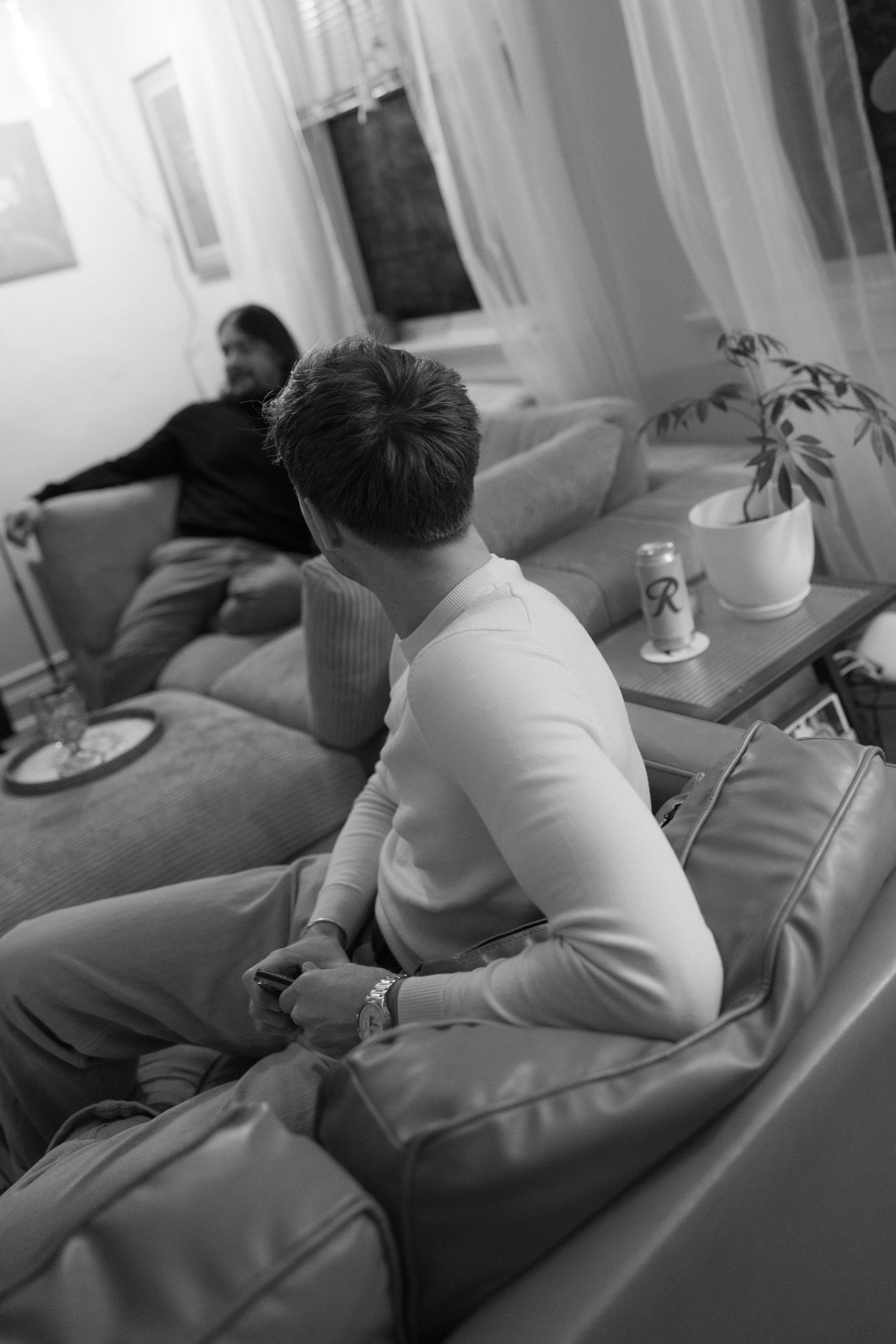 Two men in a living room, one sitting on a couch holding a phone, and the other sitting on a chair facing away from the camera. The room has a potted plant, a can of soda, and some decor on the walls.