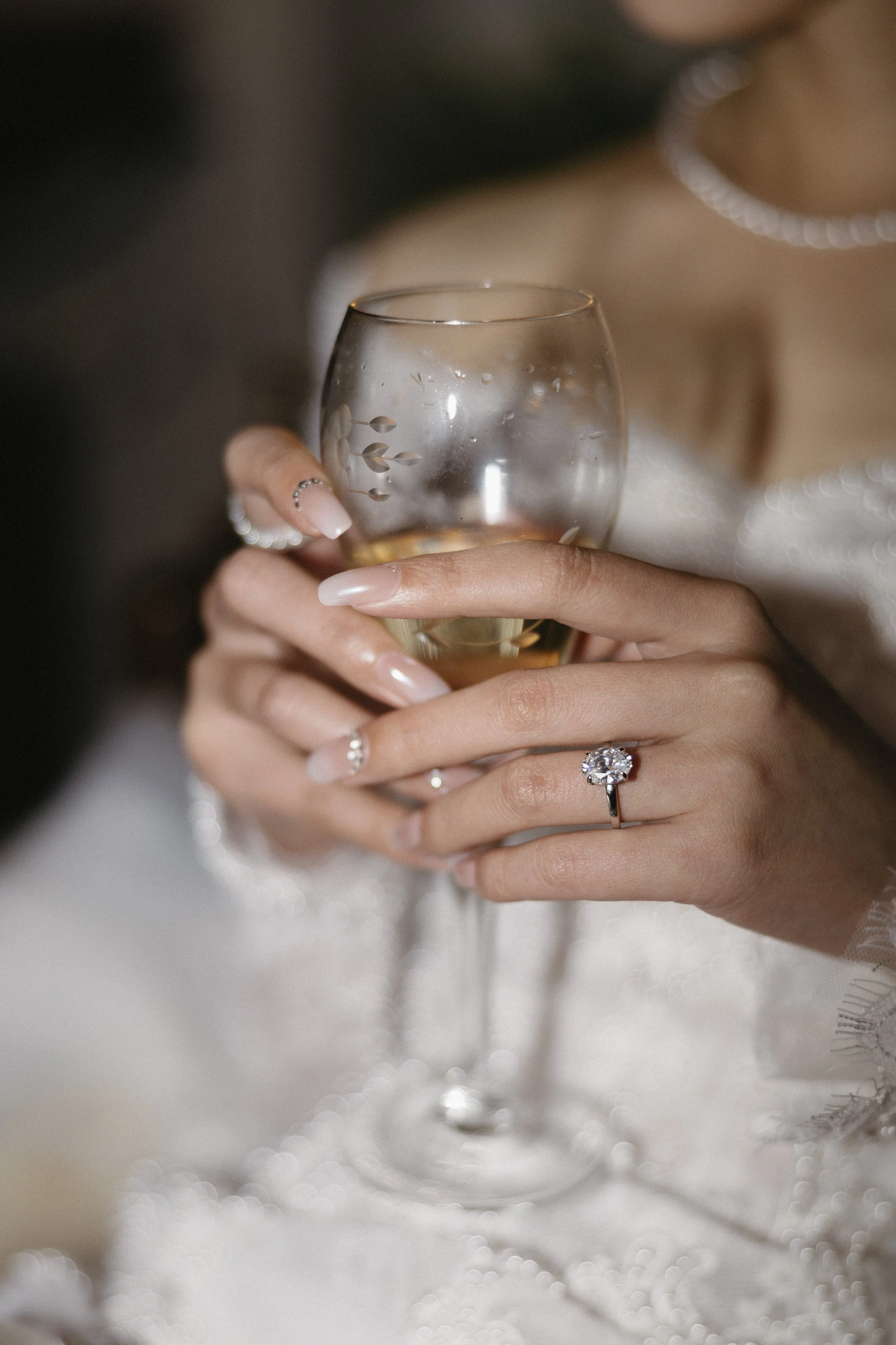 Close-up of a woman’s hand holding a glass of champagne, with a wedding ring and well-manicured nails, wearing a wedding dress with pearl jewelry visible in the background.