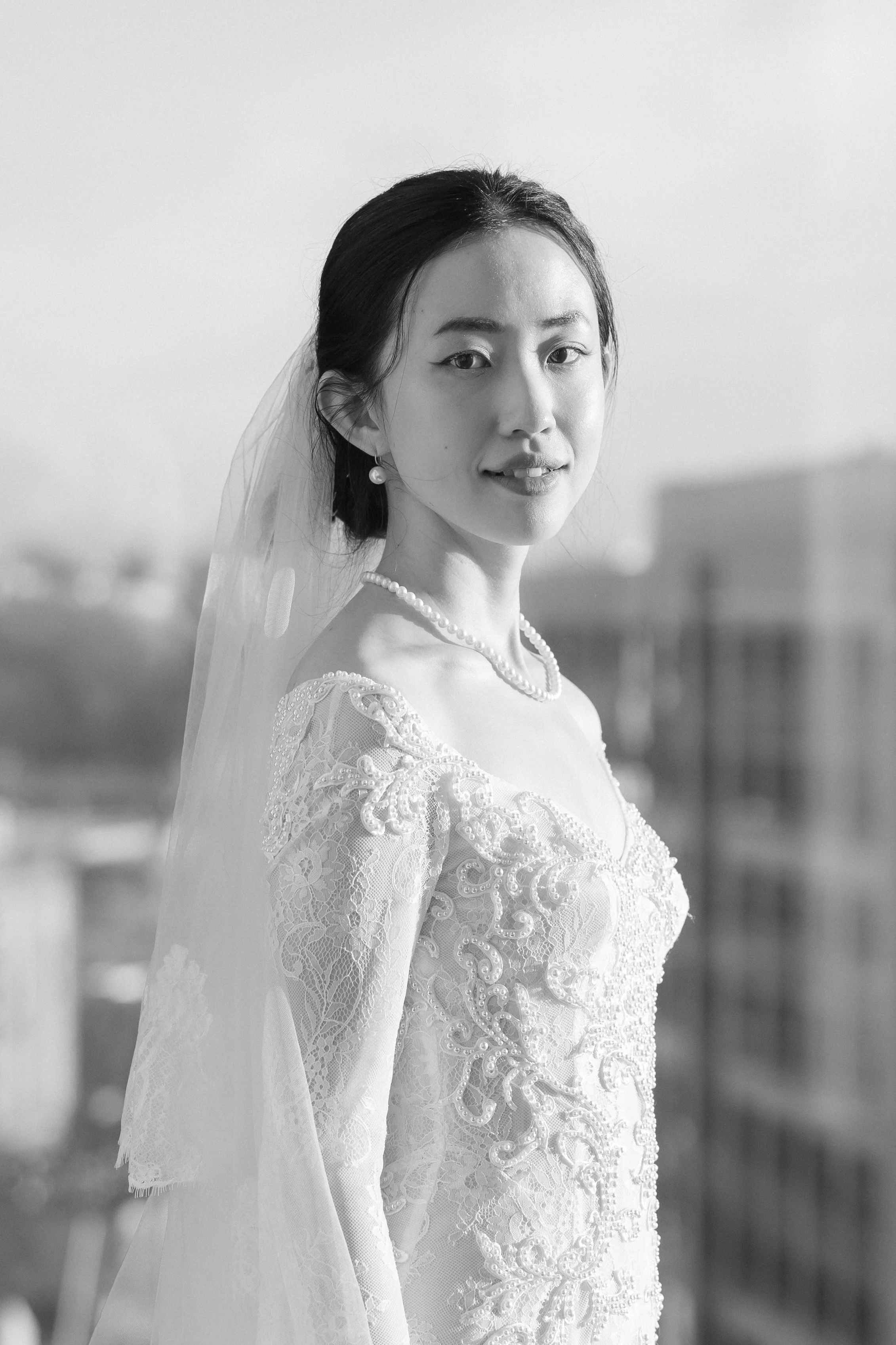 Black and white photo of a bride wearing an elegant lace wedding dress with embroidery, a pearl necklace, pearl earrings, and a sheer veil, standing outdoors with blurred cityscape background.