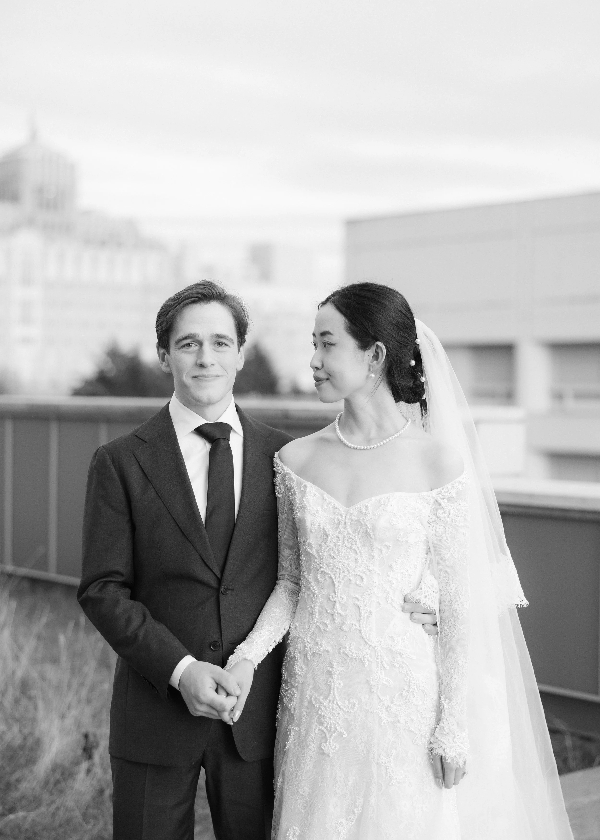 Black and white photo of a bride and groom holding hands outdoors on a city rooftop. The groom is dressed in a suit, and the bride is wearing a lace wedding gown with off-the-shoulder sleeves, pearl necklace, and veil.
