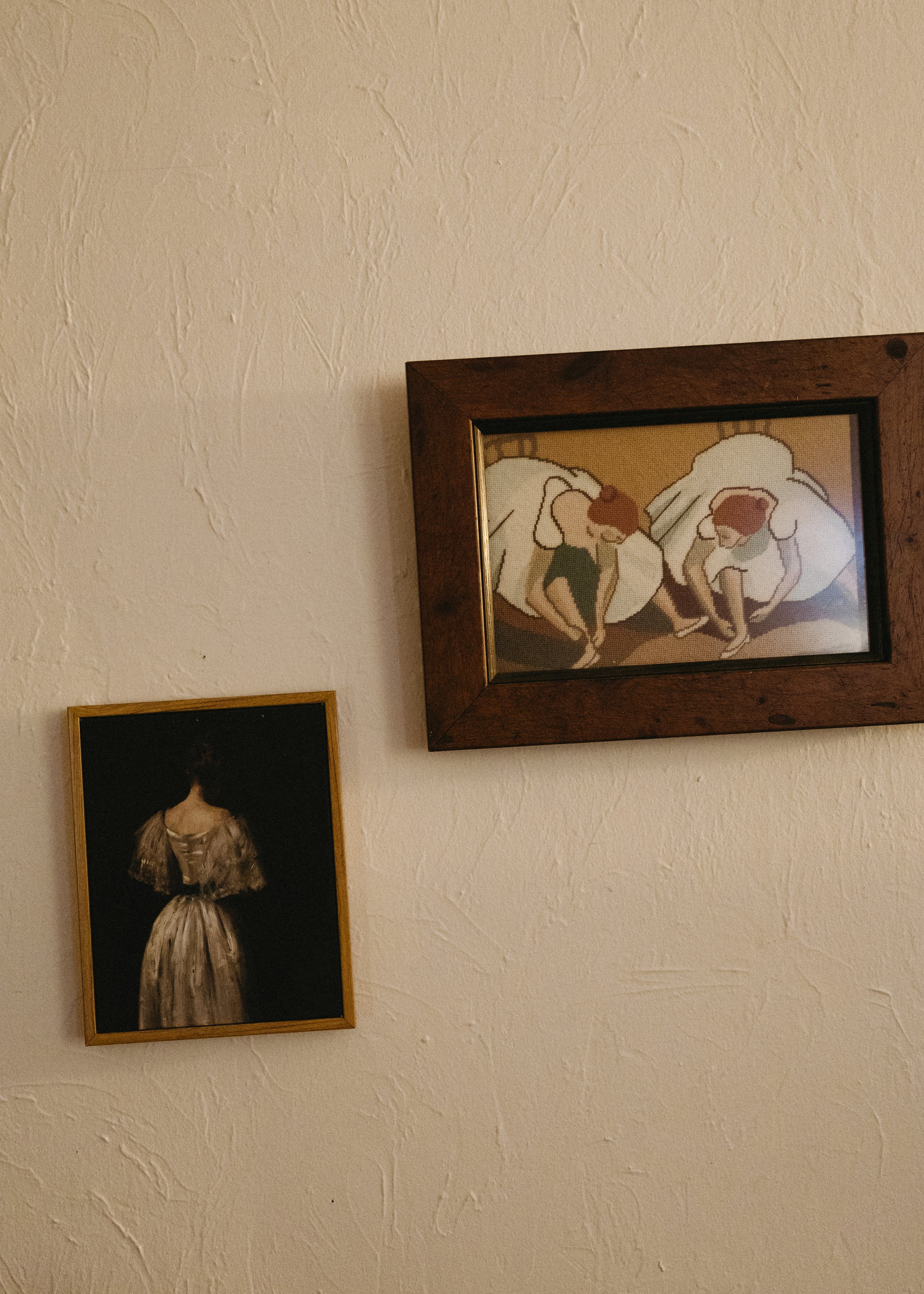 Two framed paintings on a textured beige wall. The painting on the right depicts two women with red hair in white dresses, seated with their knees bent and legs crossed, in a stylized, abstract style. The painting on the left shows the back view of a