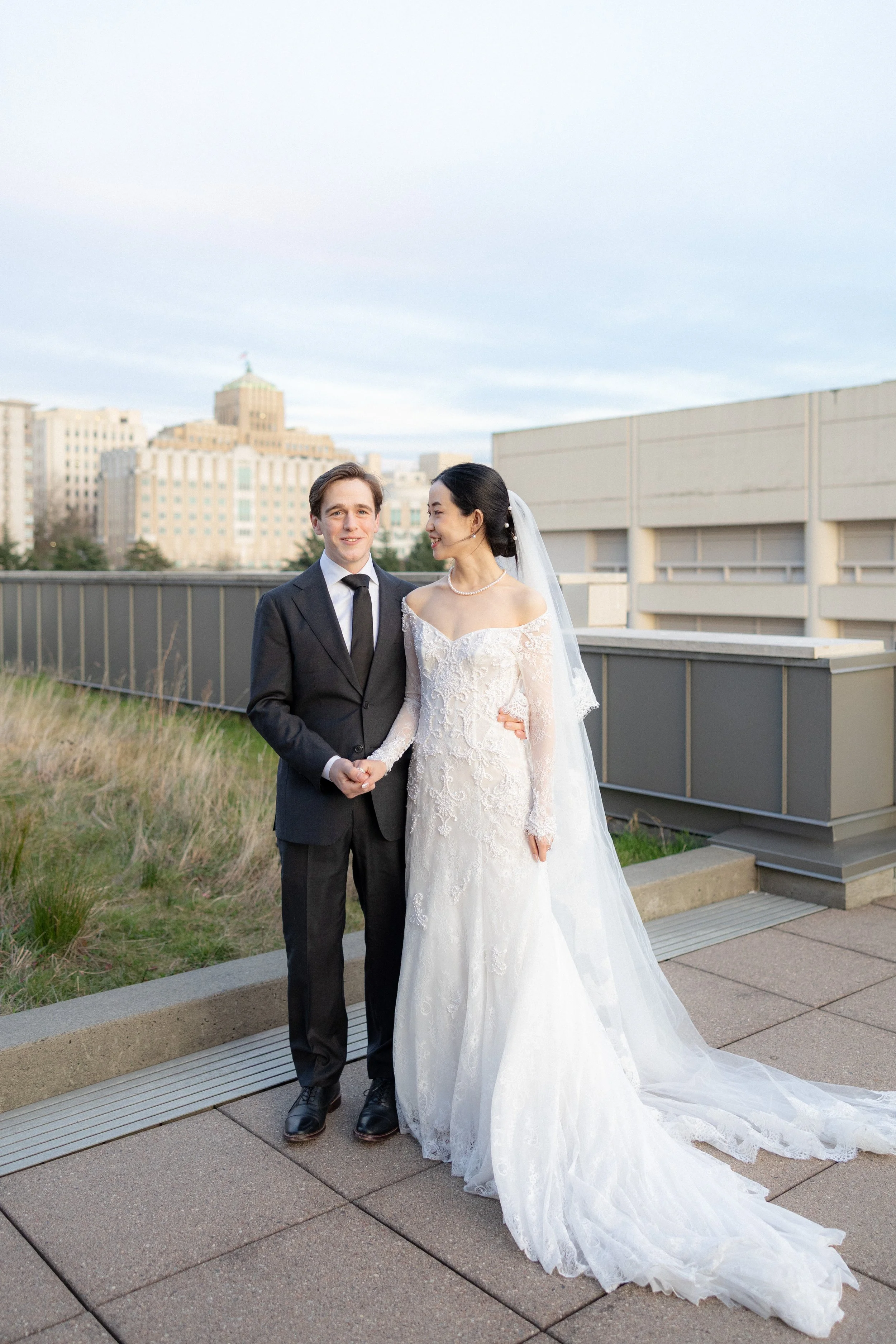 A bride and groom standing on a city rooftop, dressed in wedding attire, with the city skyline behind them during daytime.