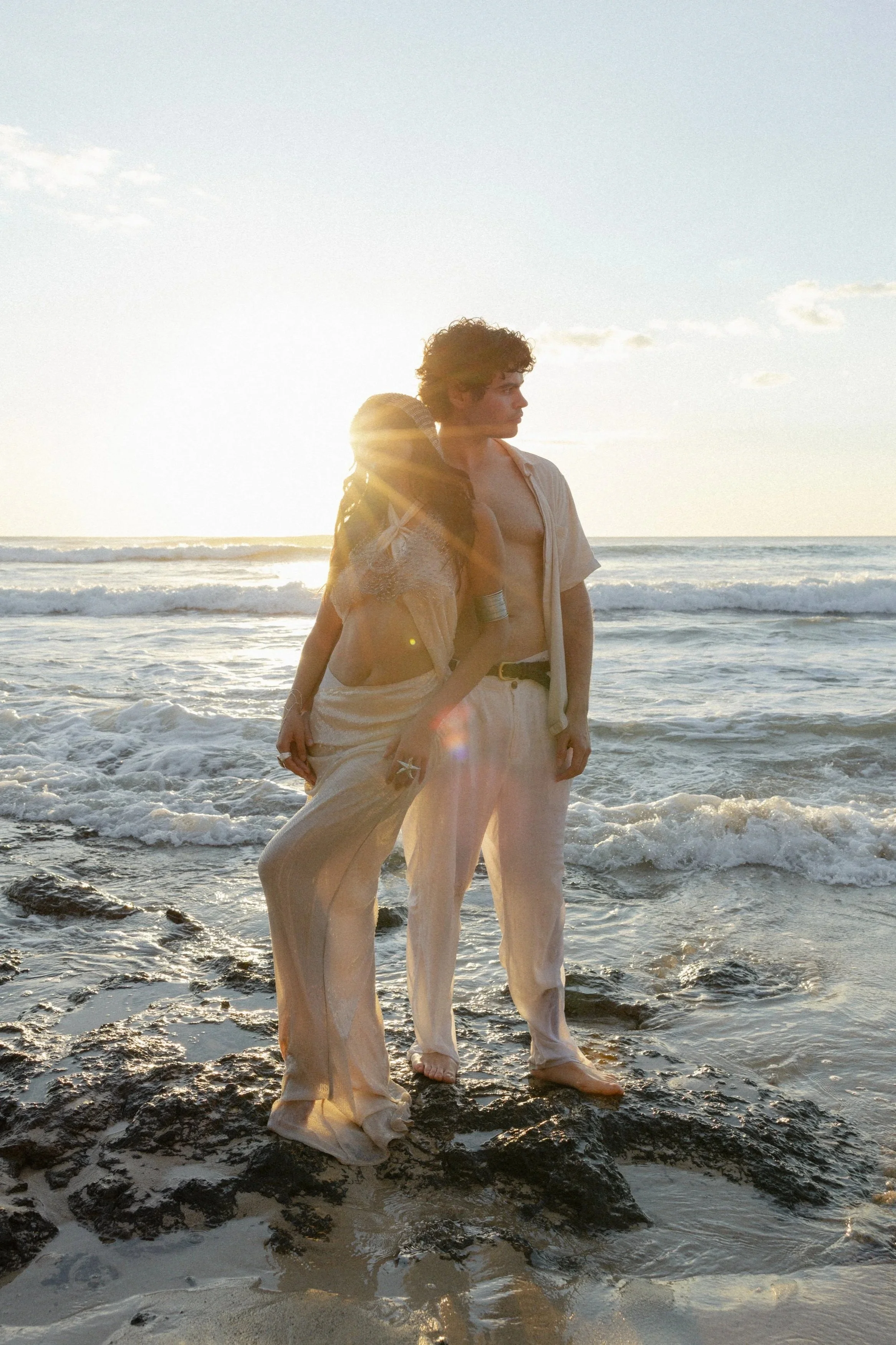Two young people, a woman and a man, standing on rocks at the beach during sunset. The woman is leaning on the man, who has his arms loosely by his sides. Both are dressed in light, flowing clothing, and the sun is setting behind them, casting a warm glow.