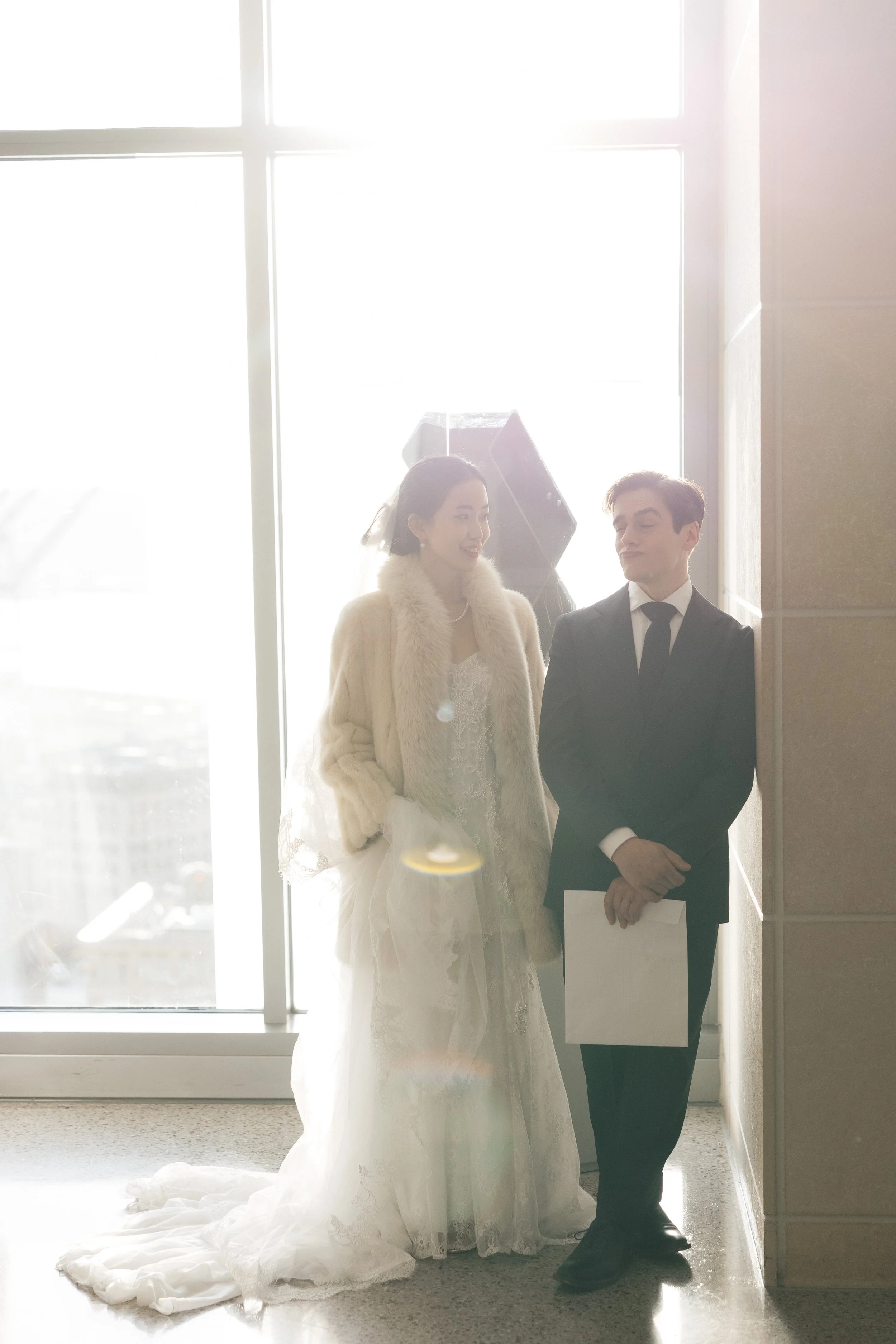 A bride and groom standing by a large window with bright sunlight. The bride is wearing a lace wedding dress, a fur coat, and a veil, and is smiling. The groom is in a black suit, white shirt, and black tie, holding a folder or book. They are in an i