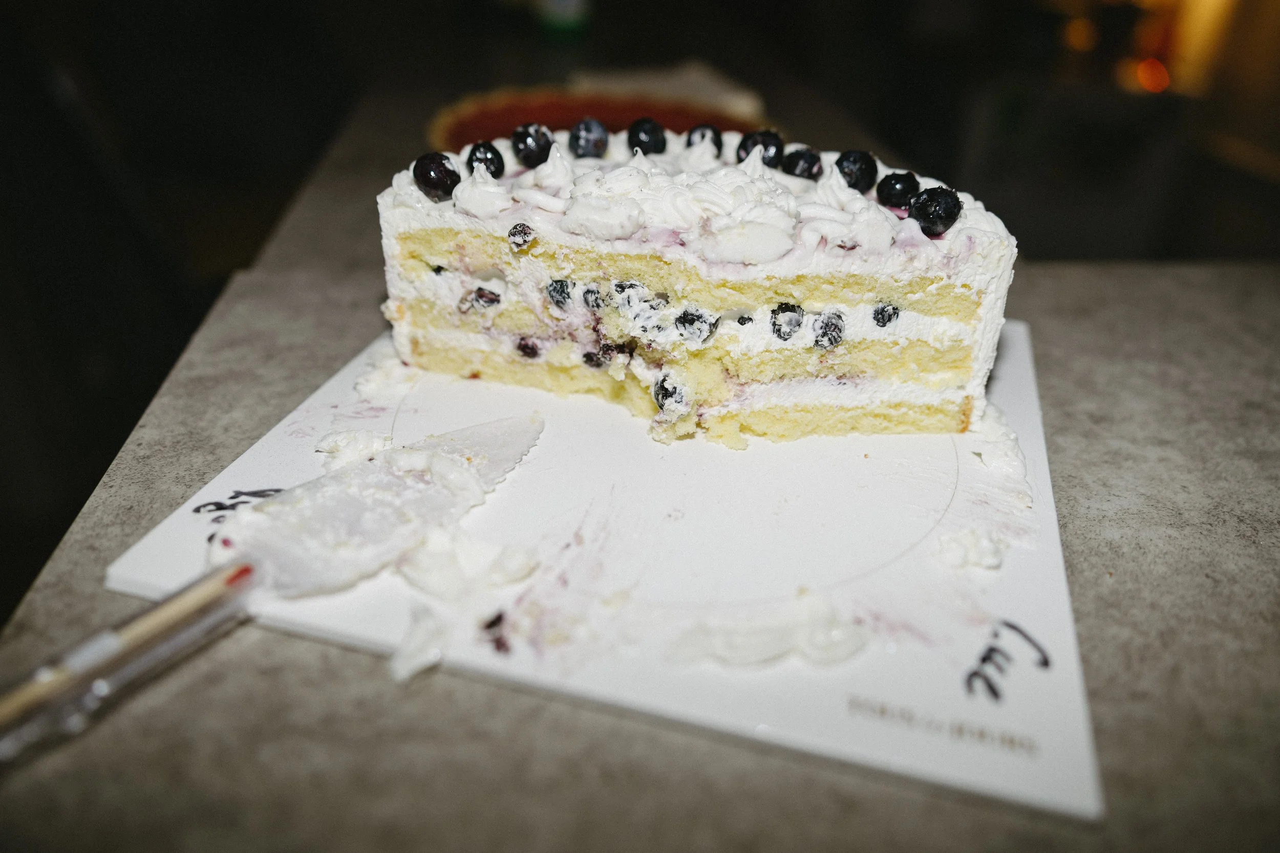 Slice of blueberry cake with white frosting and blueberries on top, on a white surface with cake remnants nearby.