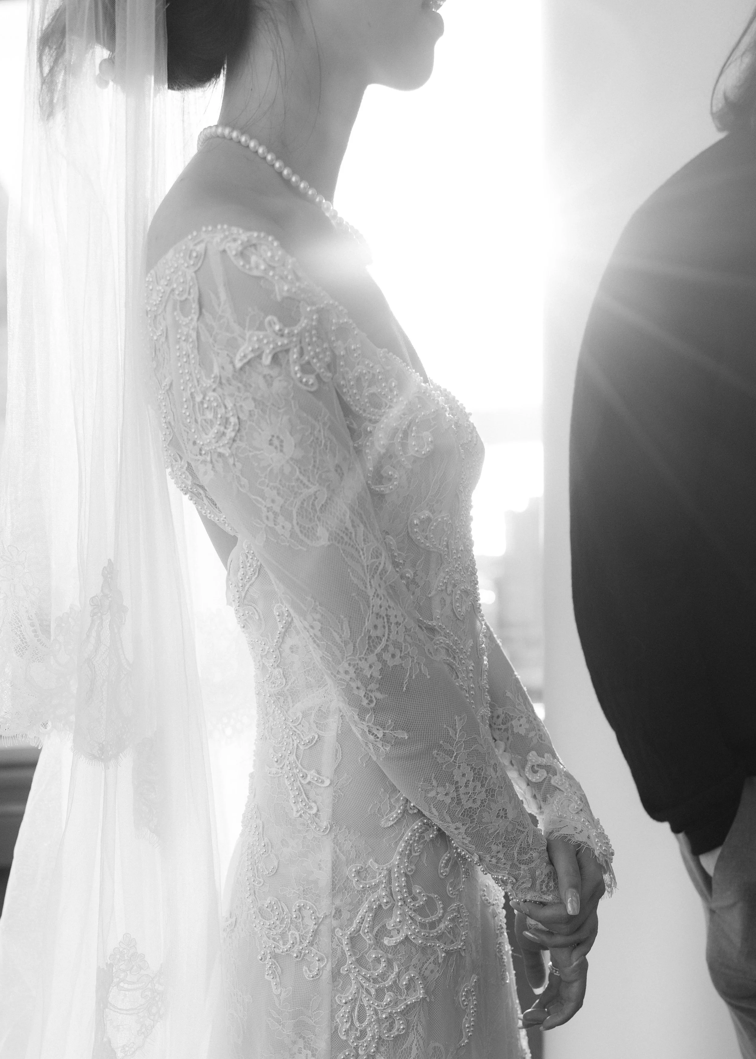 A bride in a lace wedding dress and pearl necklace holding hands with a groom dressed in a dark suit, taken in black and white.