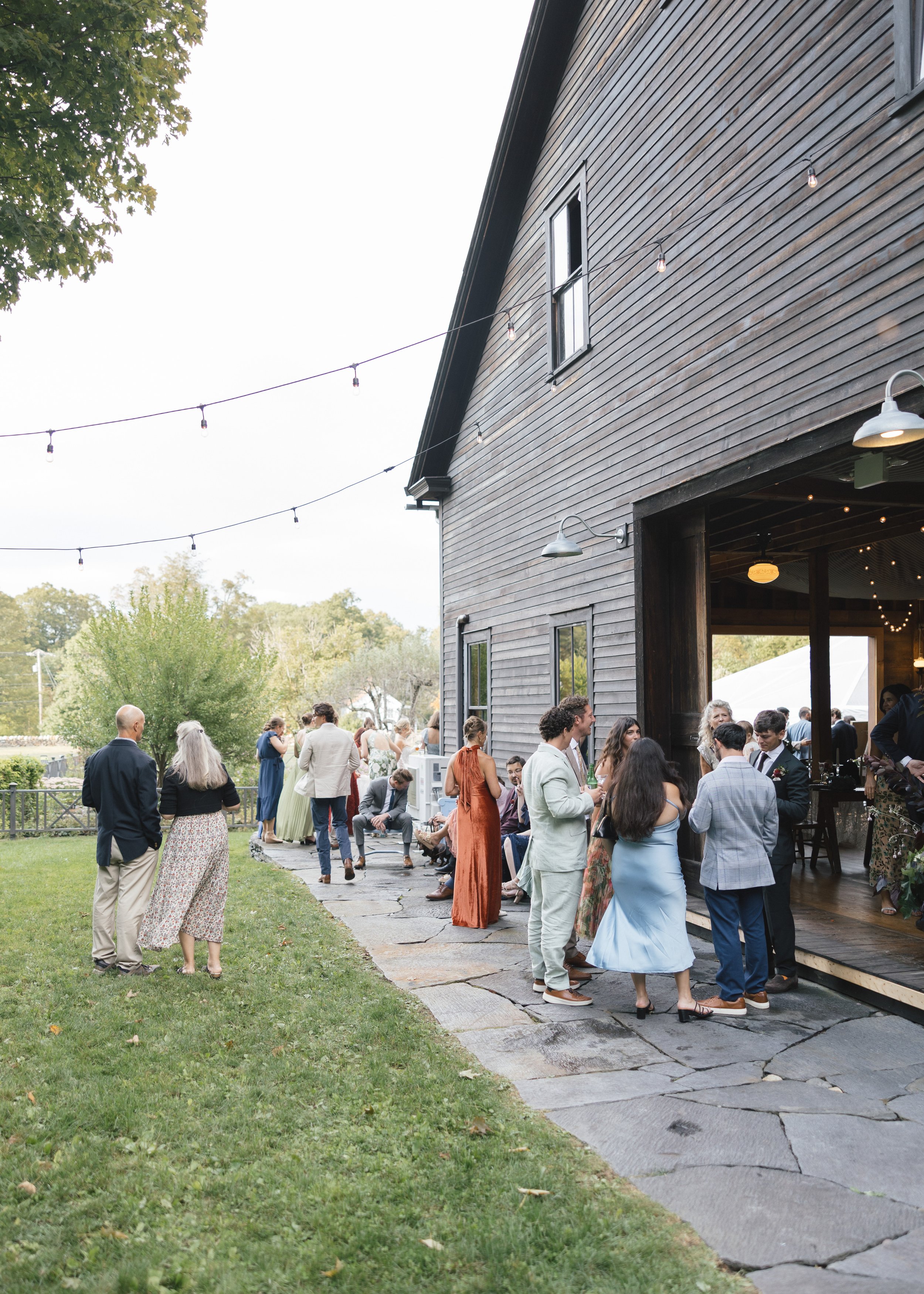 Guests at a social gathering outside a rustic barn, some socializing and others sitting on benches, on a sunny day with string lights overhead.